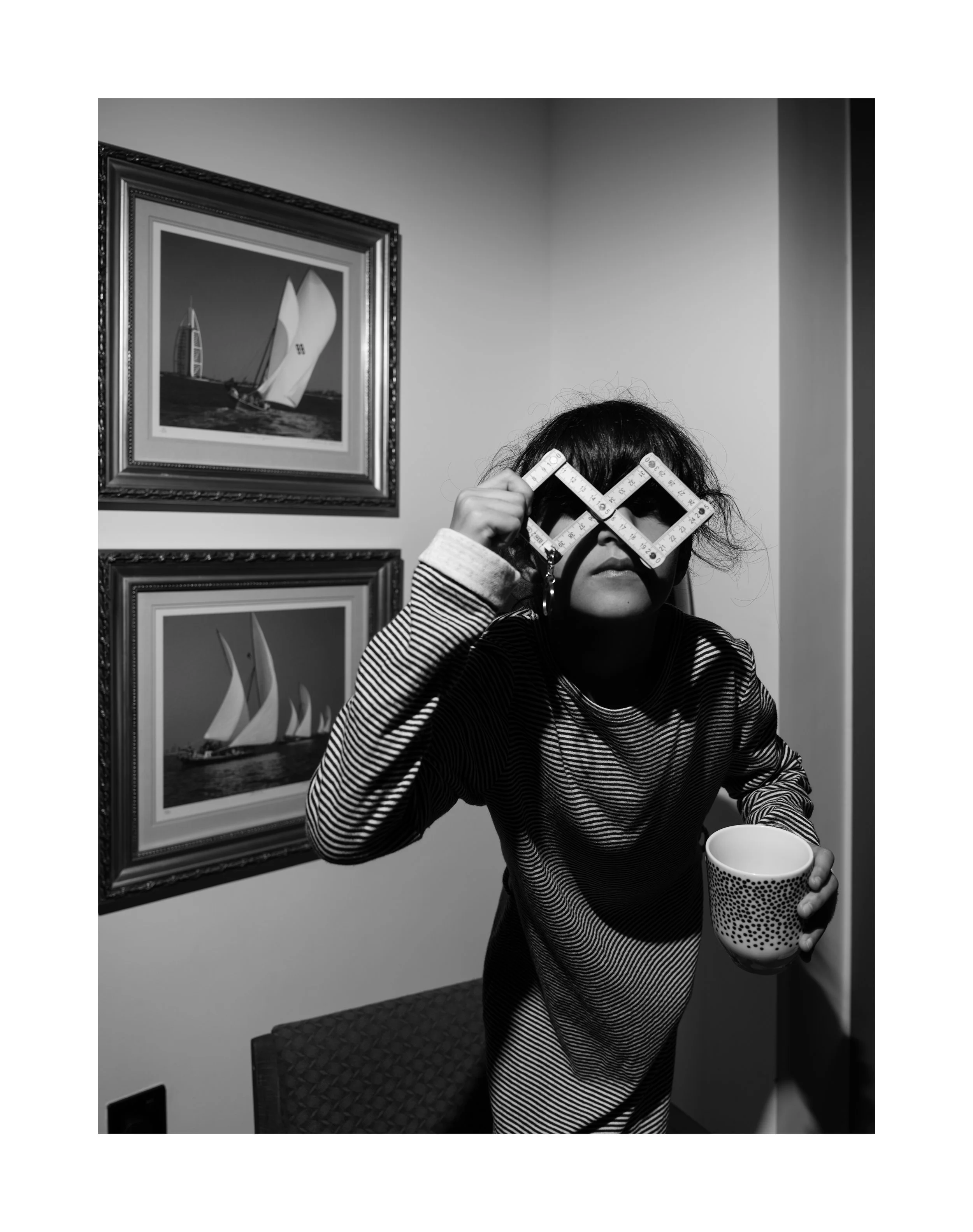 A woman with dark hair and light skin holds a mug and a geometric measuring square to her face, covering her eyes, while standing in a room with framed artwork on the wall, including a sailboat. The photo is in black and white.