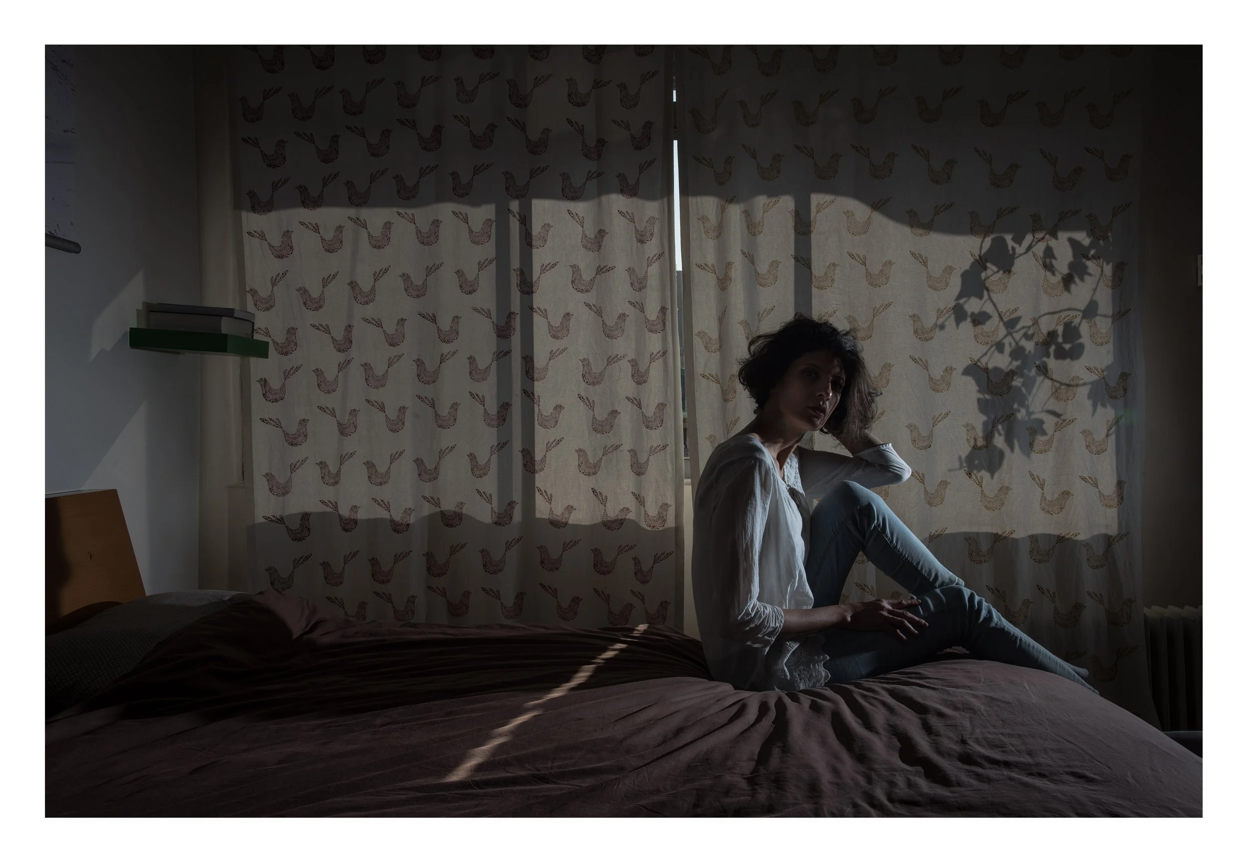 A woman with curly hair sitting on a bed in a dimly lit room with patterned curtains and shadows of a plant on the wall.