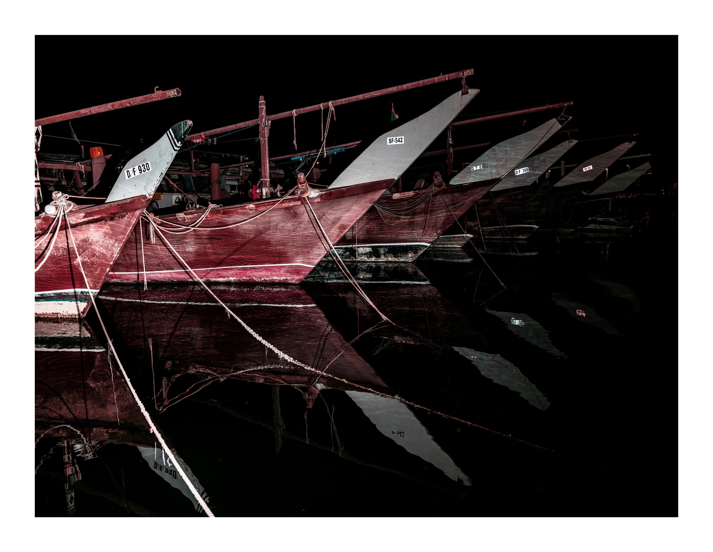 Night photograph of several sailboats docked at a marina, with hull numbers visible and their reflections on calm water.