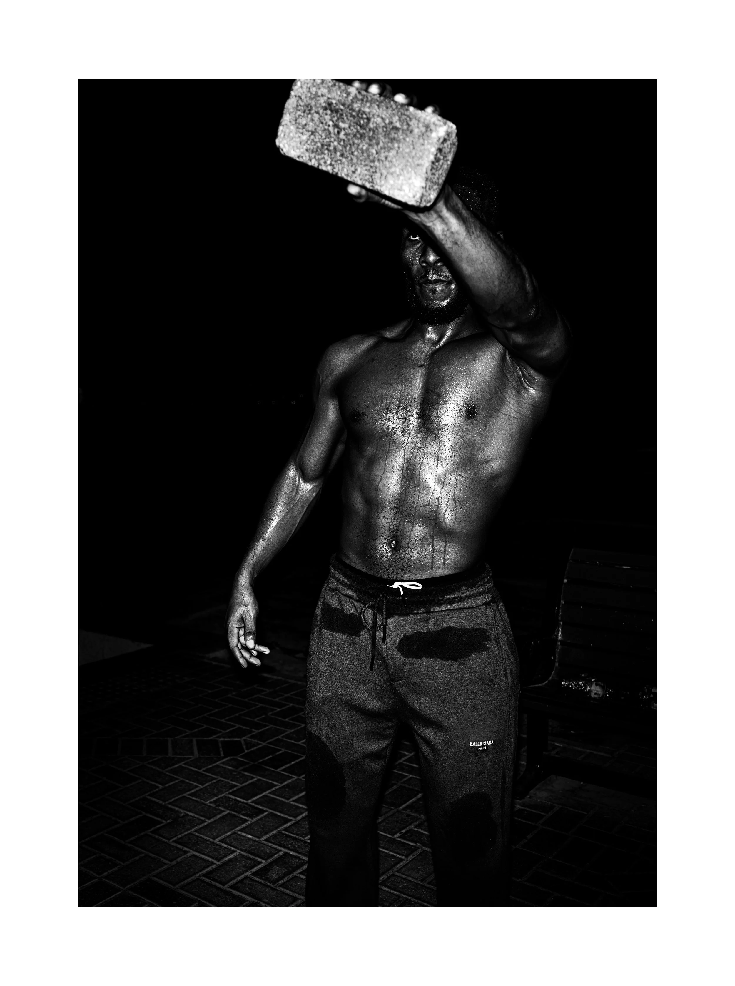 Black and white photo of a shirtless man holding a weight above his head, wearing sweatpants, standing on a brick floor indoors.