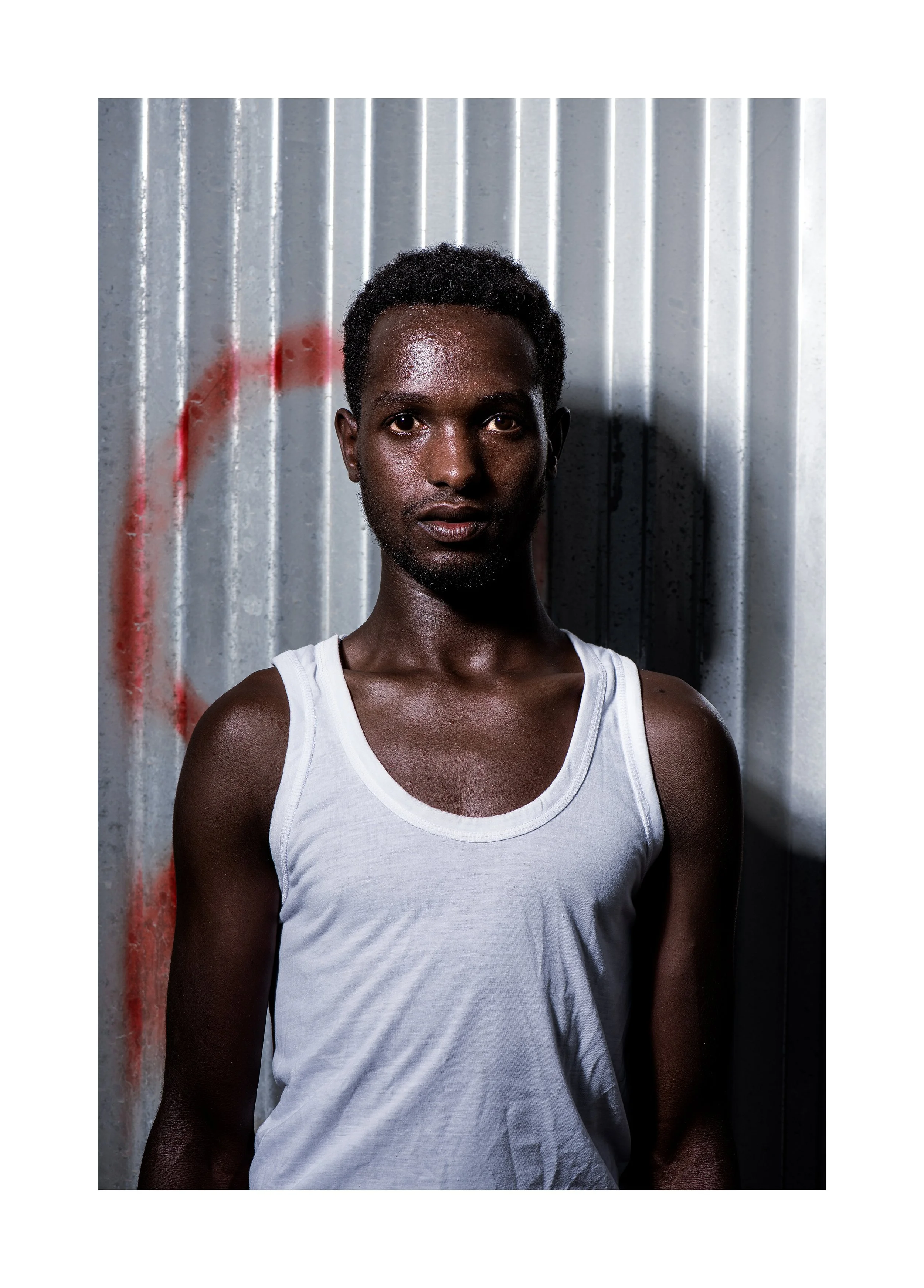 A young man with dark skin and short curly hair standing against a metallic, graffiti-marked wall, wearing a white sleeveless shirt.