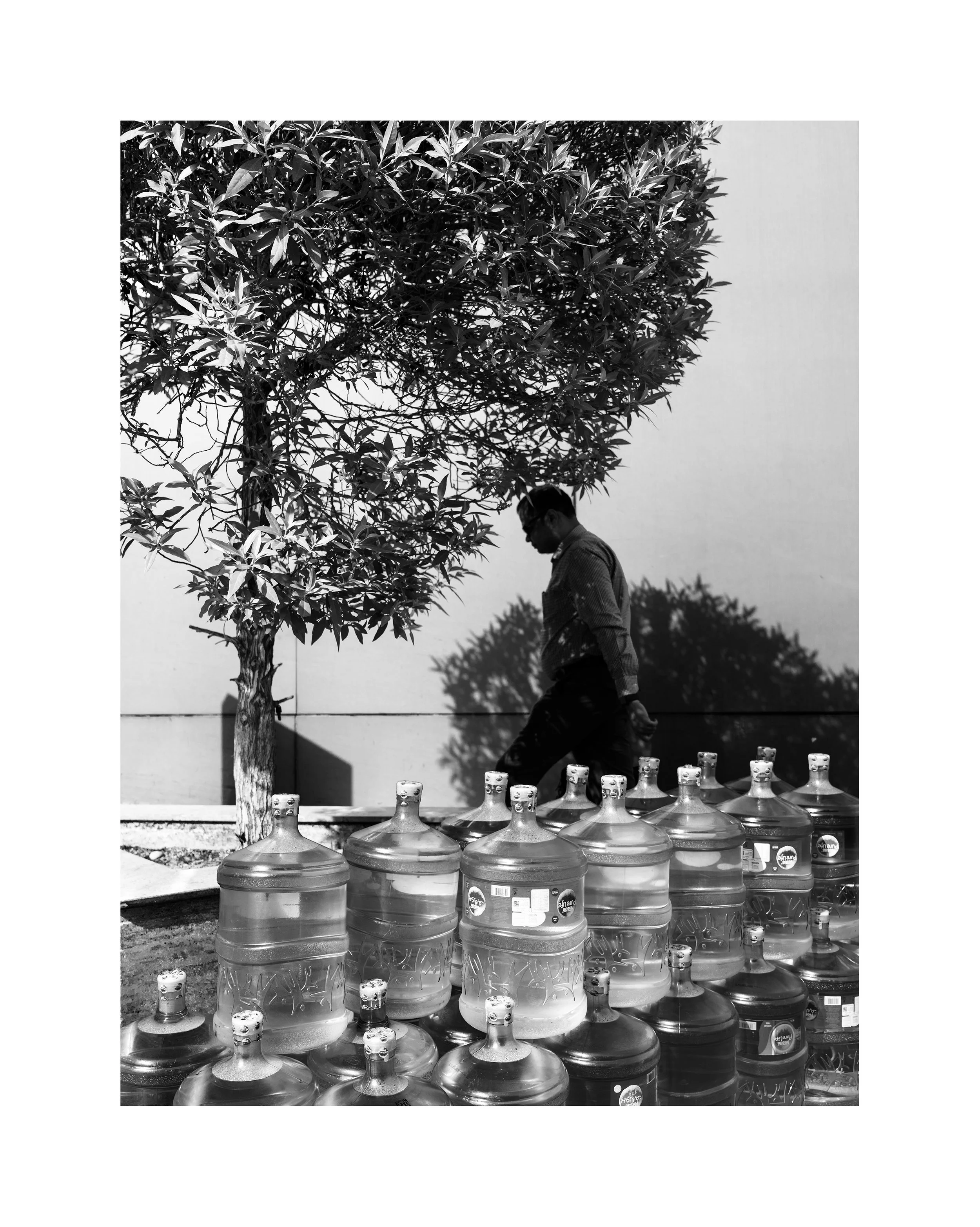 A man walking past a row of large bottled beverages outdoors on a sunny day, with a tree casting a shadow on a wall.