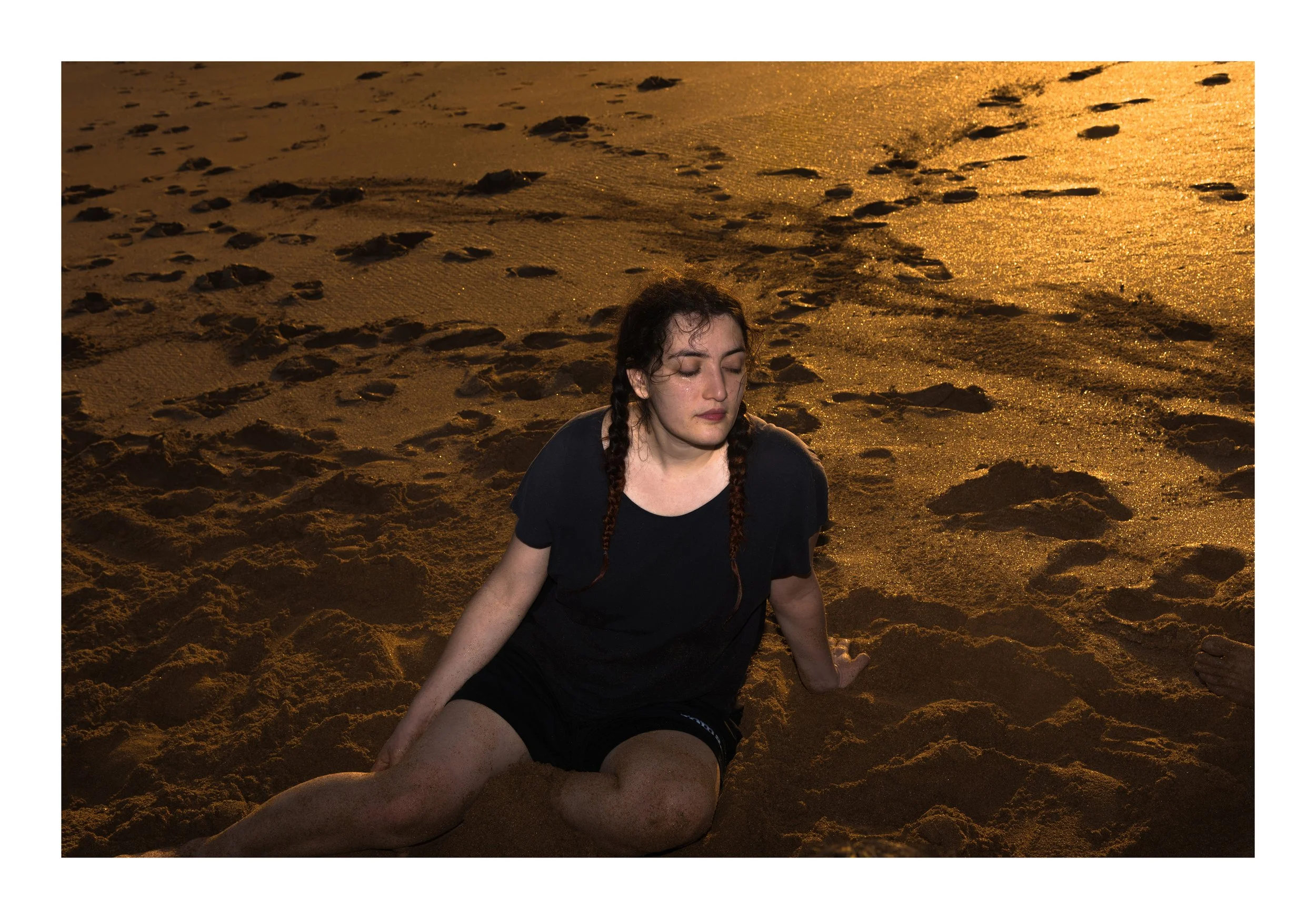 A young woman with dark hair in braids and a black outfit sitting on a sandy beach during sunset, eyes closed, with footprints in the sand around her.