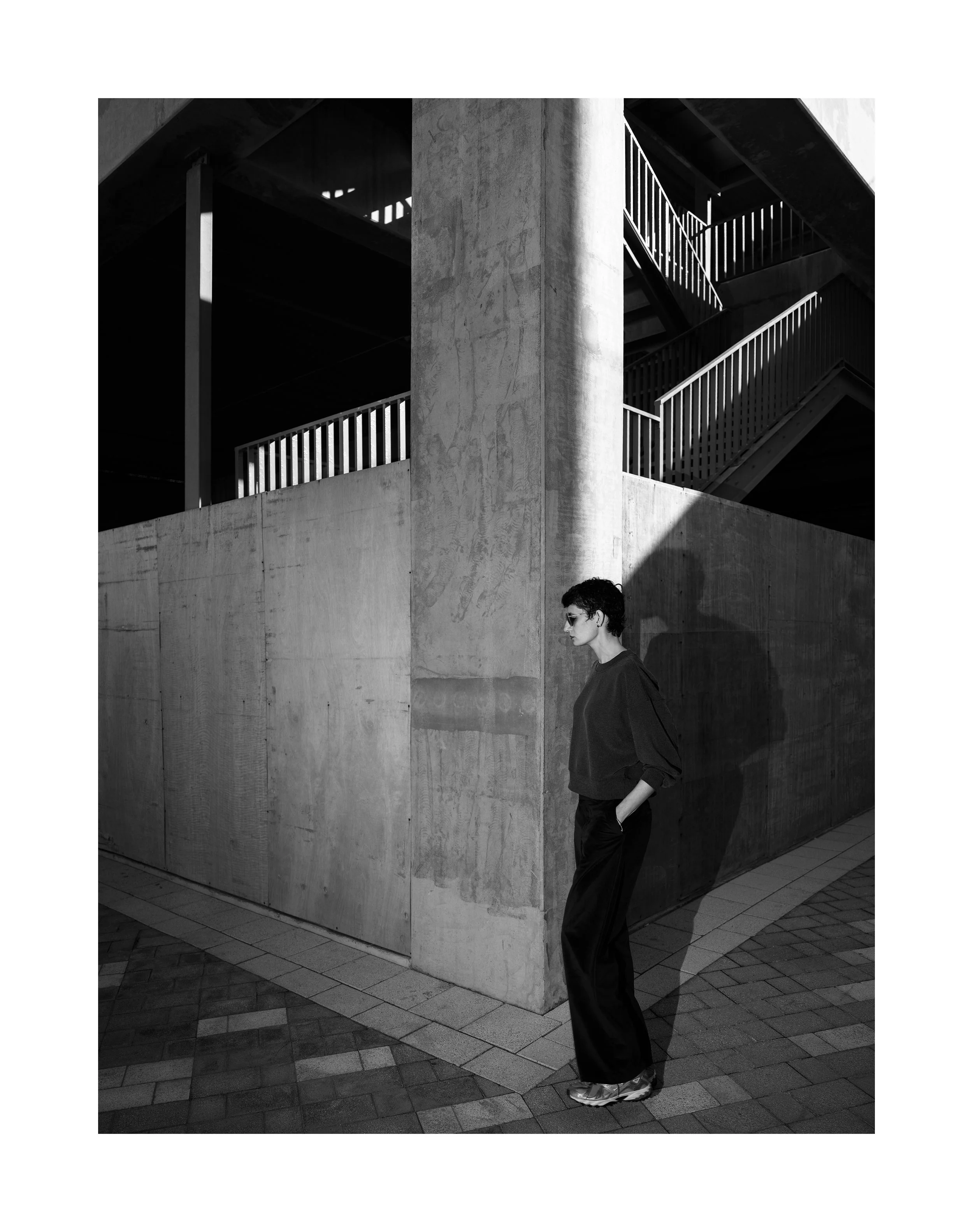 A black-and-white photo of a person standing against a concrete wall outside a modern building. The person has dark hair and sunglasses, wearing a dark oversized shirt, loose pants, and sneakers. The building has a stairway with metal railings visibl