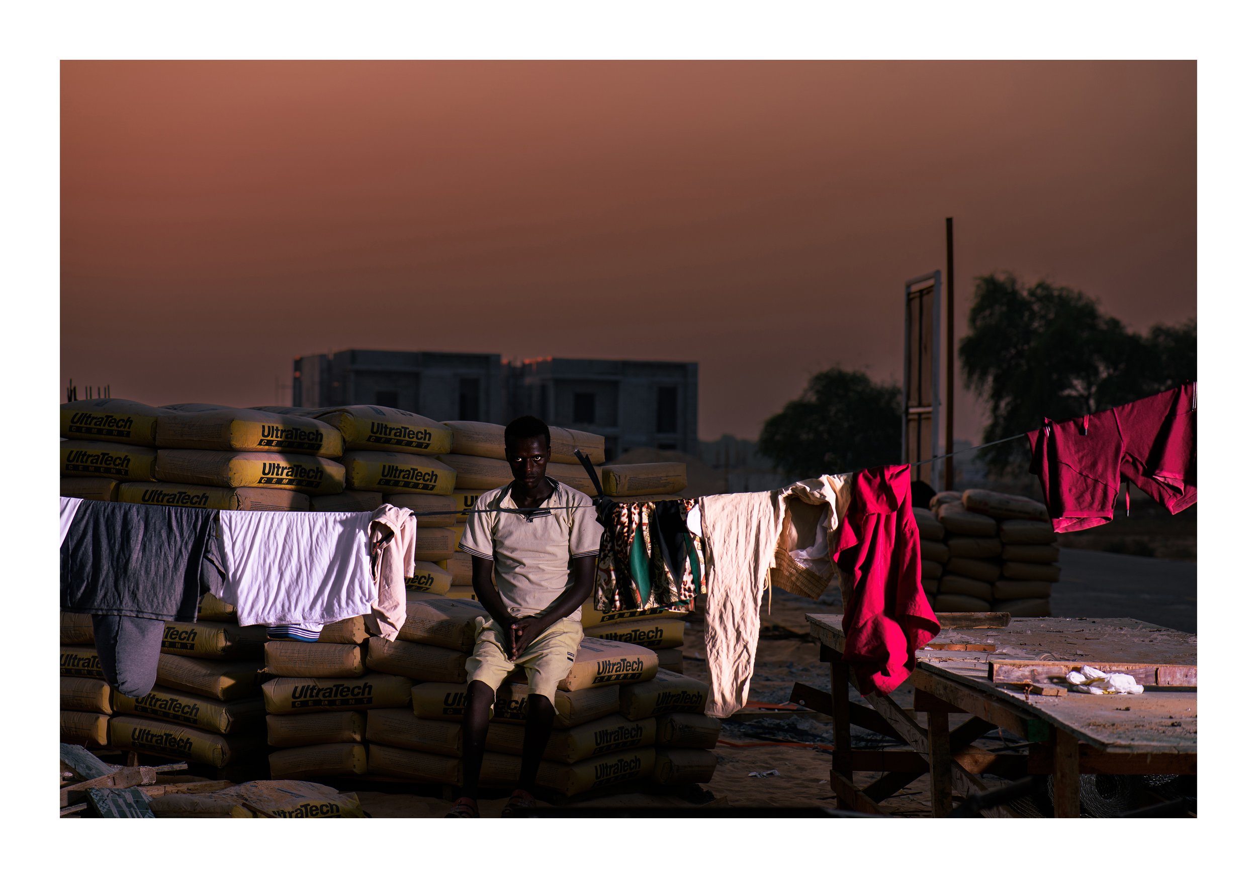 A man standing near a pile of cement bags and clothes hanging on a clothesline in an outdoor area at dusk or dawn, with buildings and trees in the background.