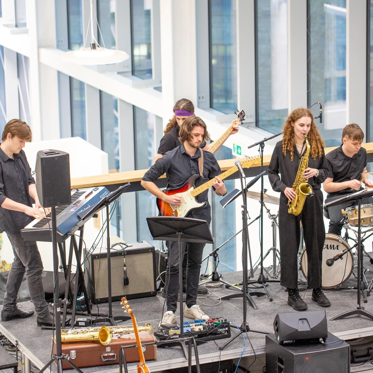 A group of five musicians performing indoors with instruments including keyboard, electric guitar, bass guitar, saxophone, and drums.