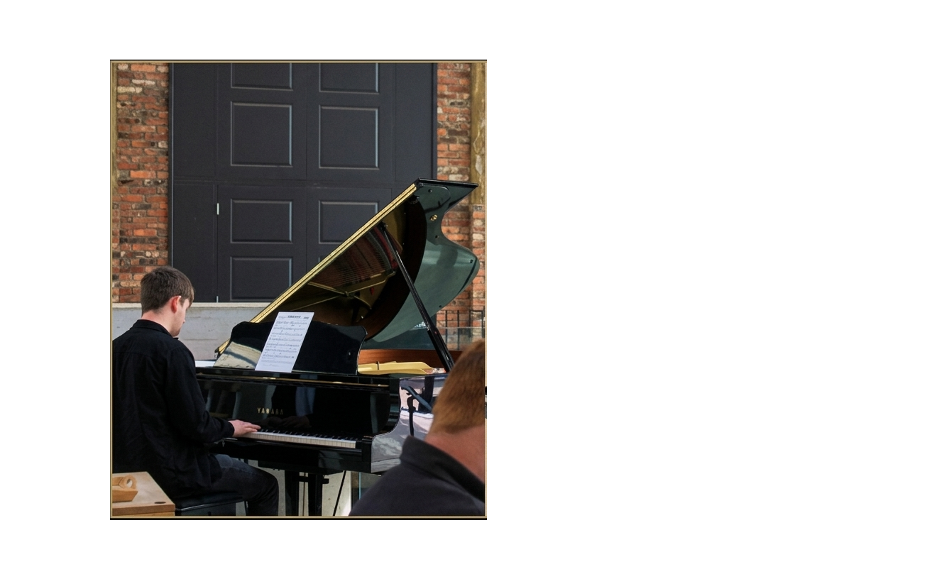 A young man playing a grand piano during a performance or practice session in a room with brick walls and a black door.