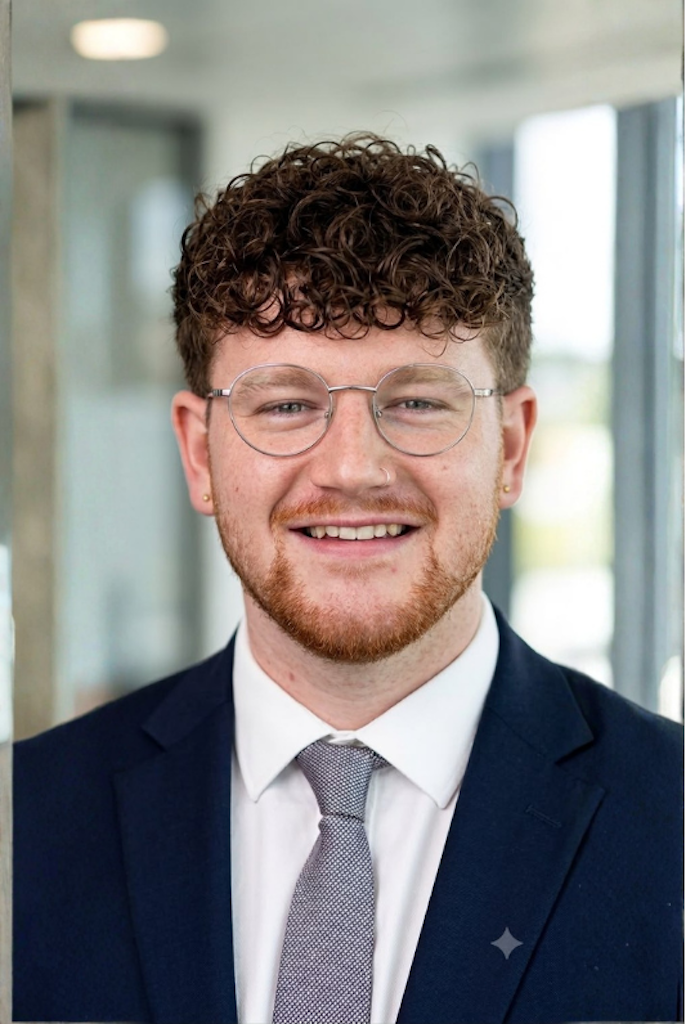 A young man with curly brown hair, glasses, and a beard, wearing a navy blue suit, white shirt, and gray tie, smiling in a modern office setting.