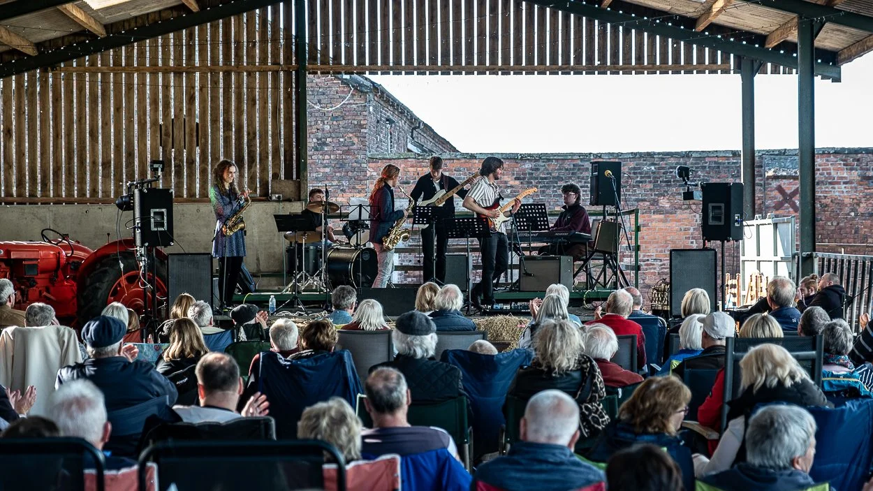 A live band performing on a rustic stage with a brick wall background, featuring vocals, guitar, keyboard, drums, and saxophone, in front of an audience seated outdoors under a wooden pavilion.