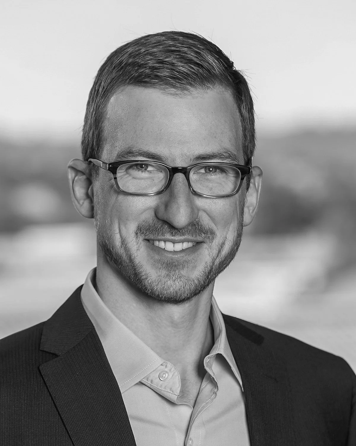 Black and white portrait of a smiling man wearing glasses, a suit jacket, and a collared shirt, with a blurred outdoor background.