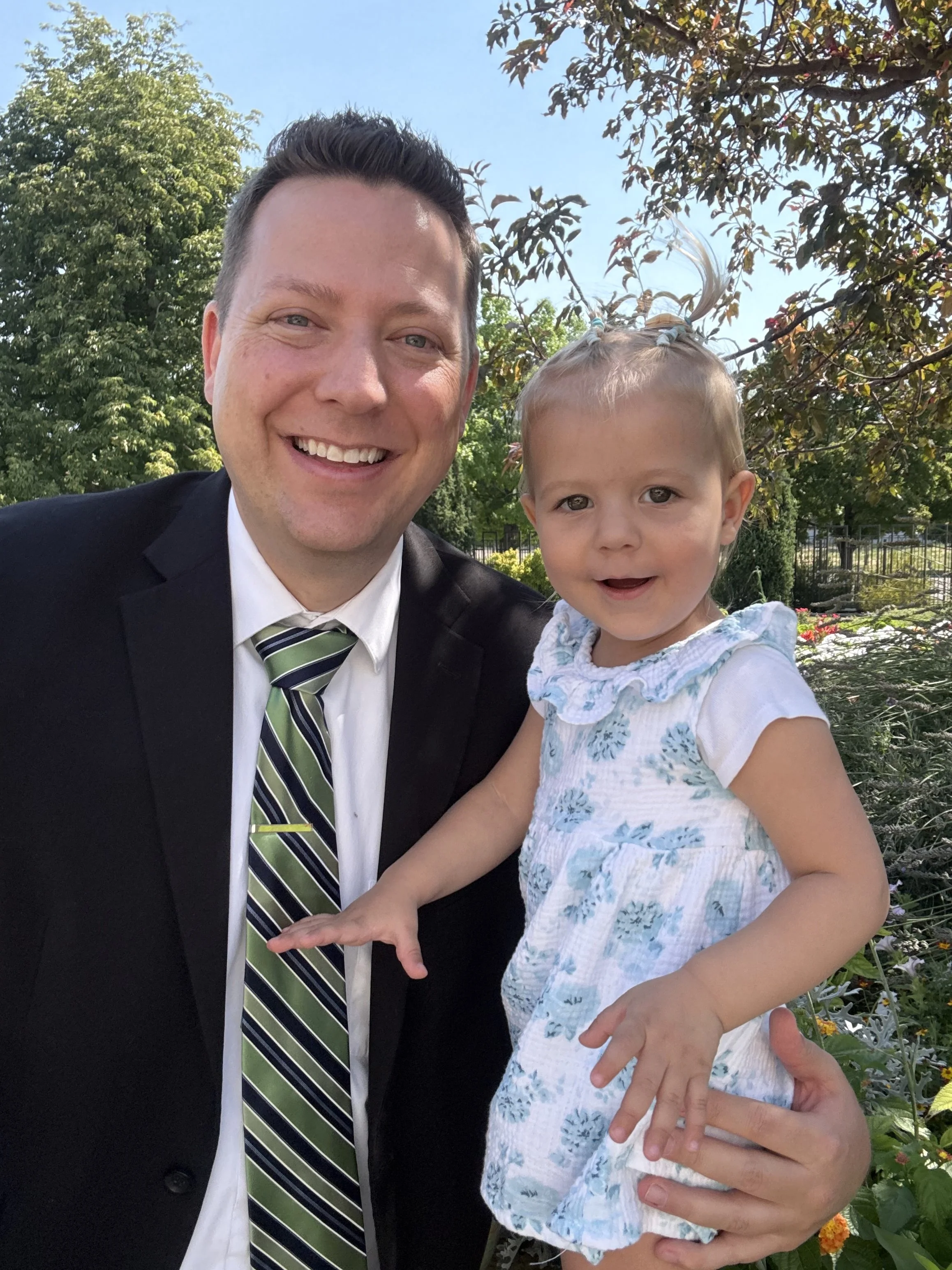 A smiling man in a black suit, white shirt, and green striped tie, holding a young girl with blonde hair styled in small ponytails and wearing a white dress with blue floral patterns, outdoors on a sunny day with green trees and colorful flowers in the background.