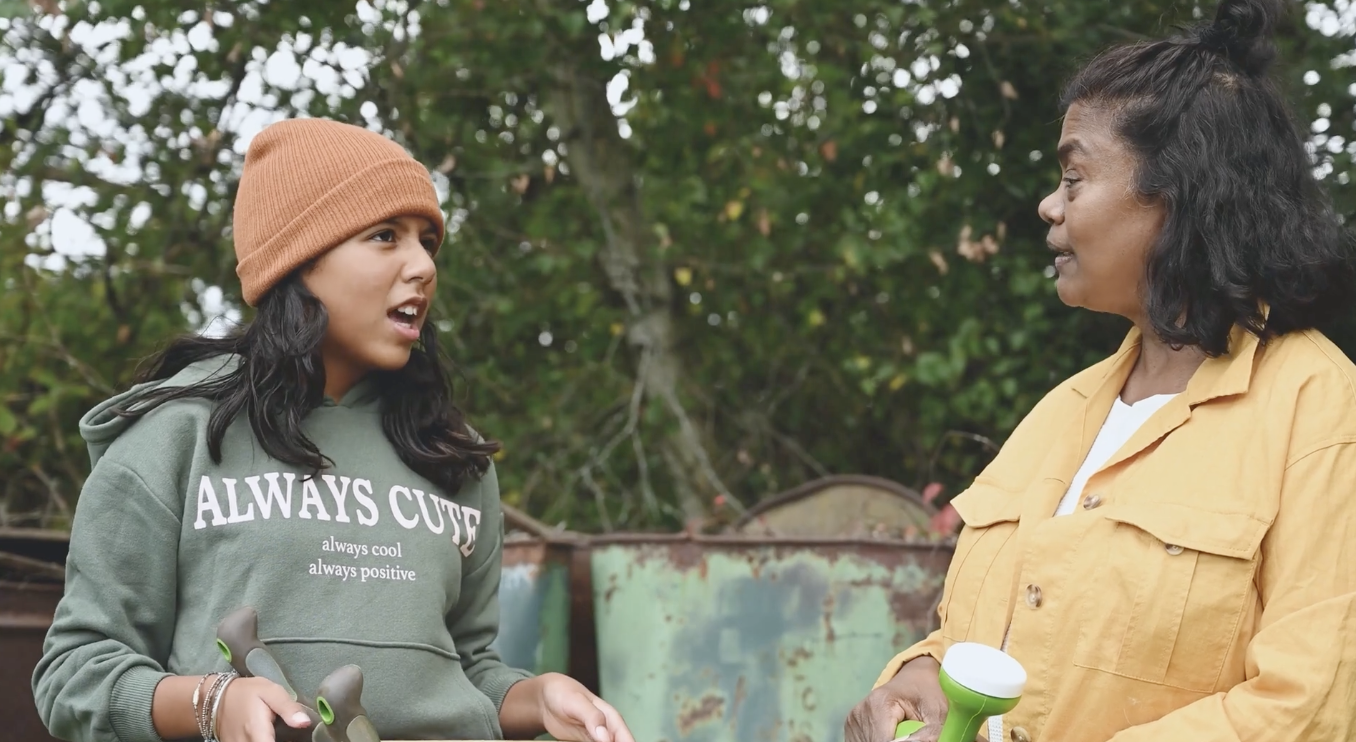 A young woman and an older woman are having a conversation outdoors, with green bushes and trees in the background. The young woman is wearing a brown beanie and a green hoodie that says 'ALWAYS CUTE,' while holding a broom. The older woman is wearing a yellow jacket and also holding a tool.