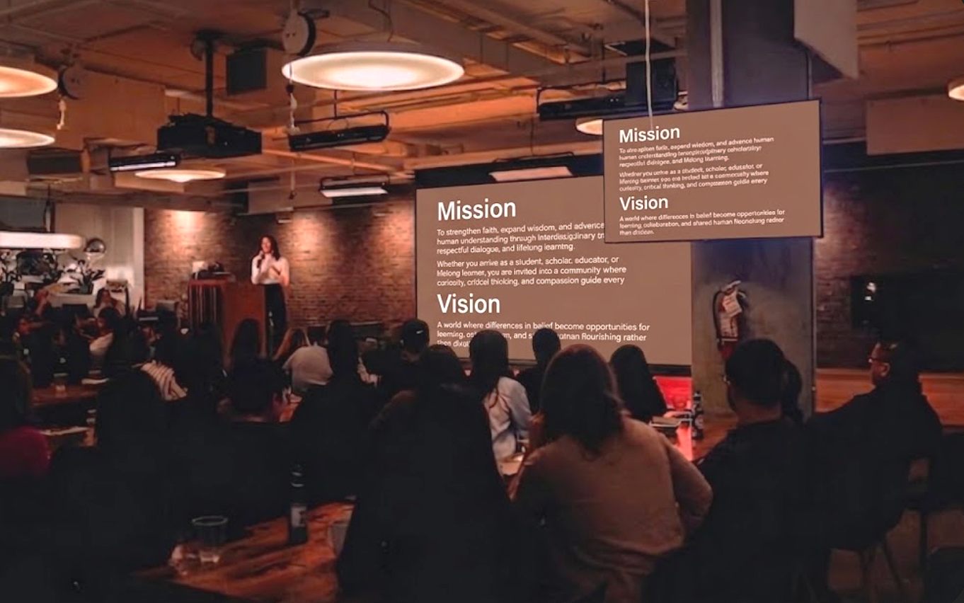 A woman giving a presentation in a dimly lit room with a brick wall background and large screens displaying mission and vision statements, while an audience listens.