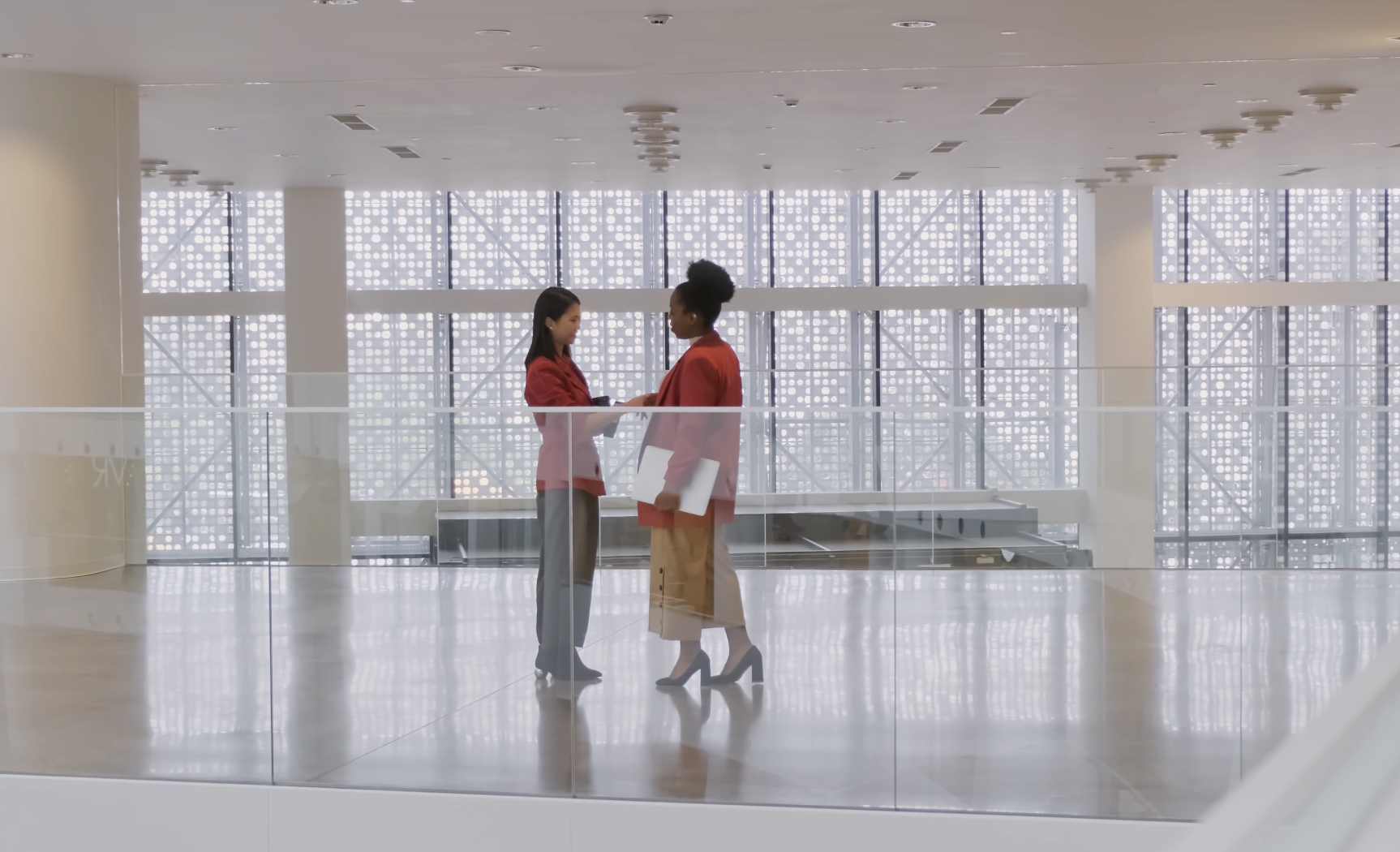 Two women, dressed in business attire, are standing and having a conversation in a modern, brightly-lit office space with a glass railing and large patterned windows.