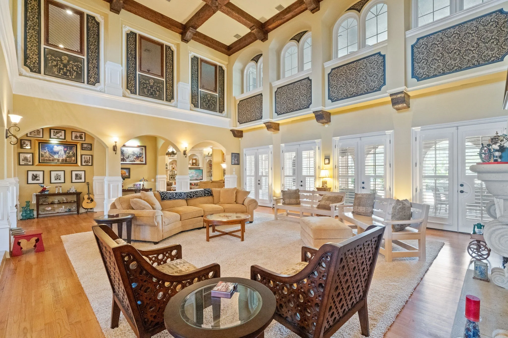 Living room with high ceilings, large windows, and a balcony. It has beige sofas, wooden chairs with decorative backs, a guitar, bookshelves, tables, framed pictures on the wall, and various decorative items. The flooring is wooden with a large area 