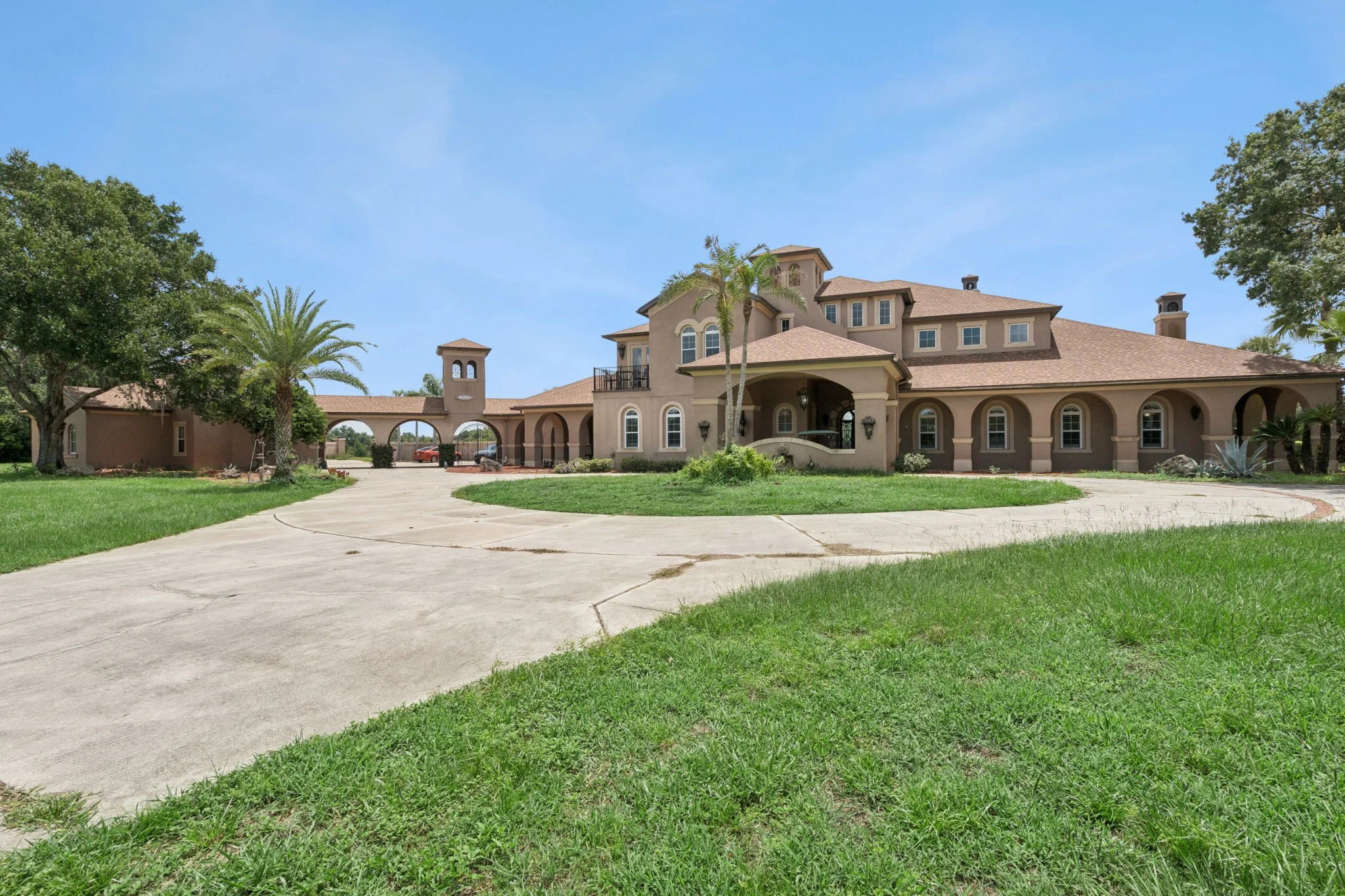 Large beige multi-story house with arched doorways, palm trees, a driveway, and lush green lawn under a blue sky.
