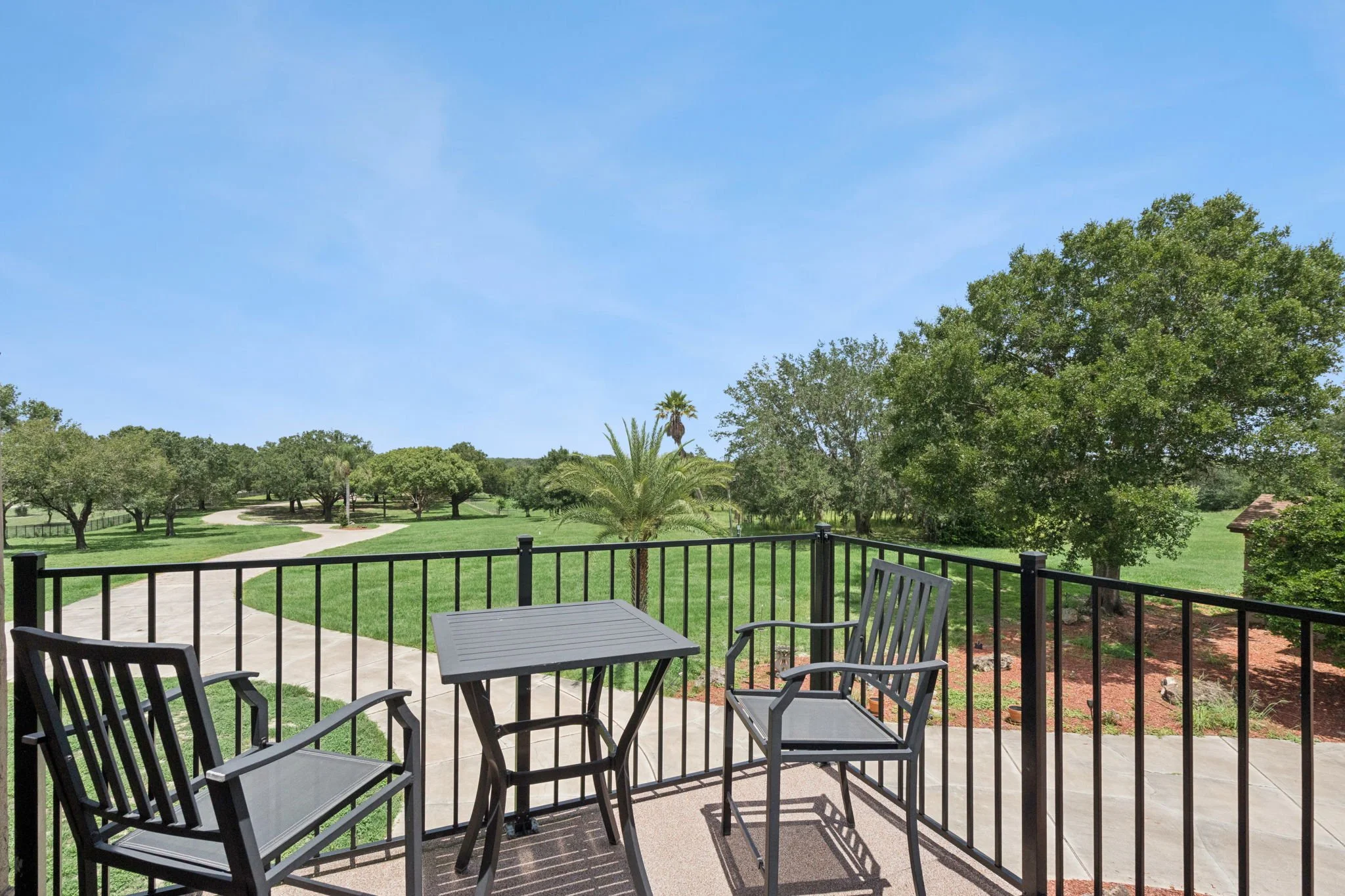 View from a patio with black metal table and chairs overlooking a lush green park with trees, pathway, and a blue sky with some clouds.
