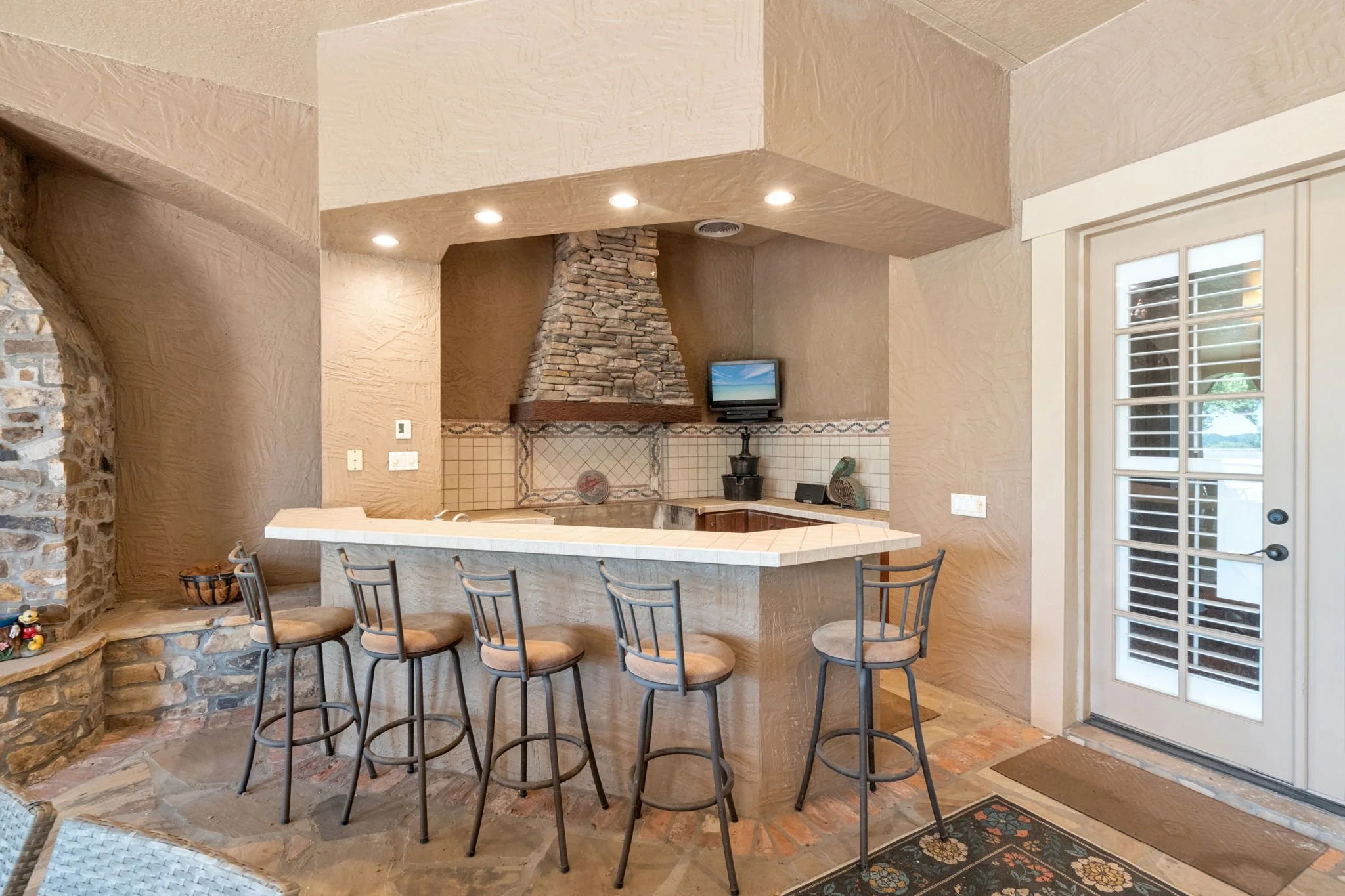 A kitchen with a beige textured wall and cabinets, a stone fireplace with a small TV, and a curved kitchen counter with six chairs. There is a sliding glass door with white shutters and a decorative rug on the floor.