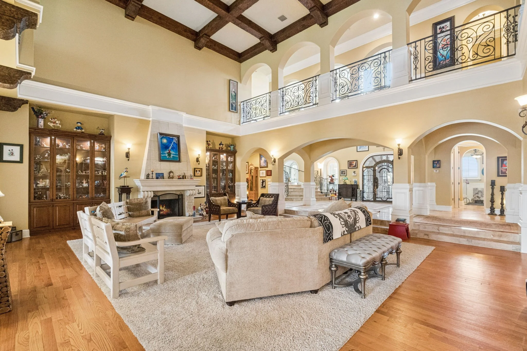 Spacious living room with beige walls, wood flooring, and high ceiling with exposed wooden beams. Features a cream-colored sectional sofa, armchairs, and a white wooden side table. Decorated with framed artwork, collectibles, and a fireplace. Large d