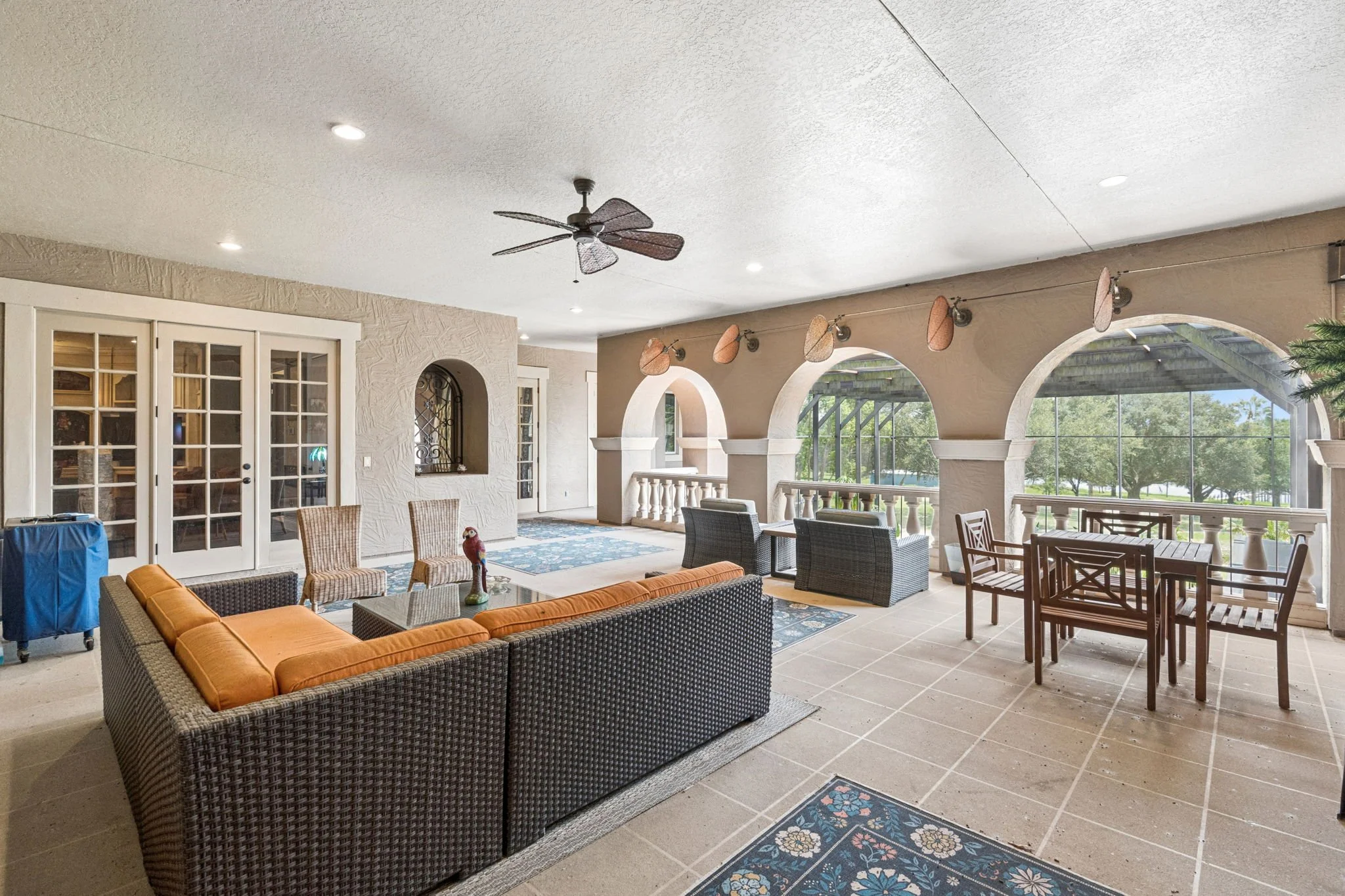 A covered outdoor patio area with wicker and wooden furniture, a ceiling fan, decorative wall hanging, and large arched openings with views of trees and an outdoor ceiling covered area.
