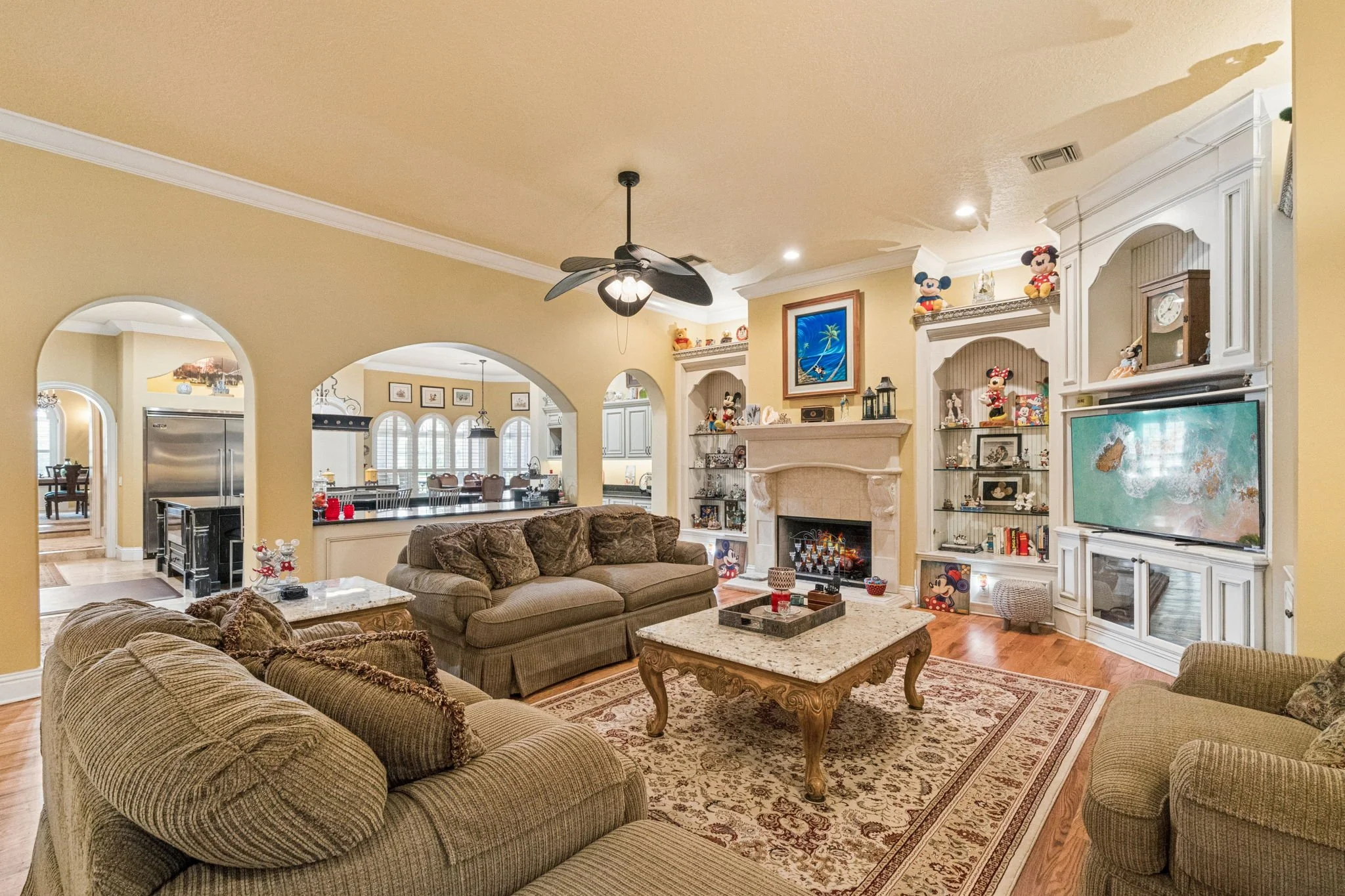 Living room with beige walls, a ceiling fan, a fireplace with decorative items, a large television, and Mickey Mouse figurines on shelves and furniture.