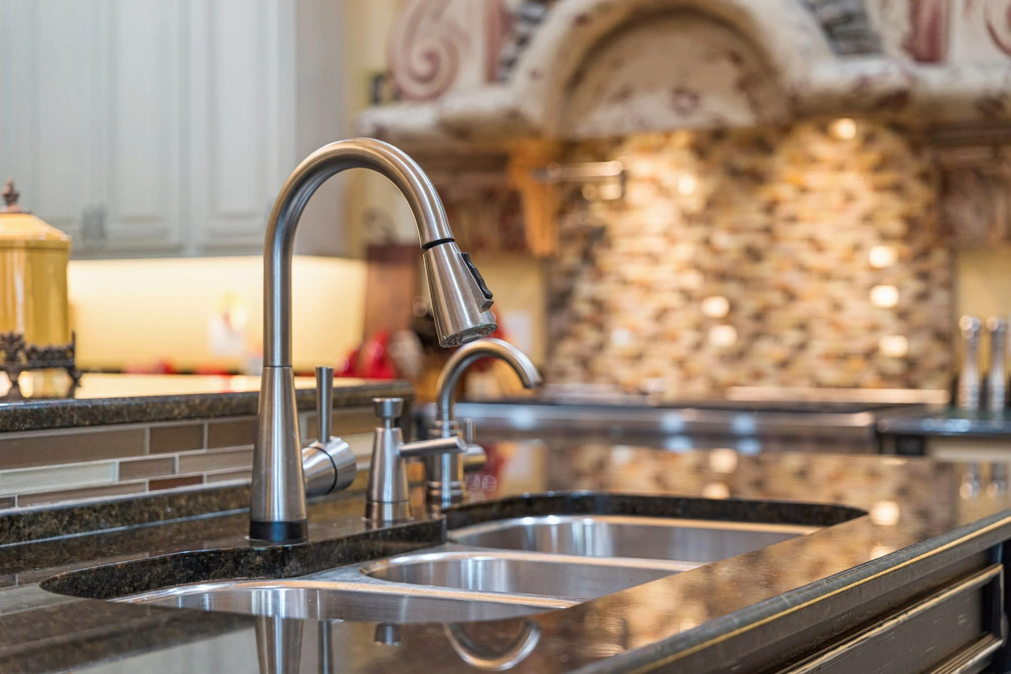 A kitchen sink with stainless steel faucets, a granite countertop, and a decorative tiled backsplash, with a blurred brick wall and arch detail in the background.