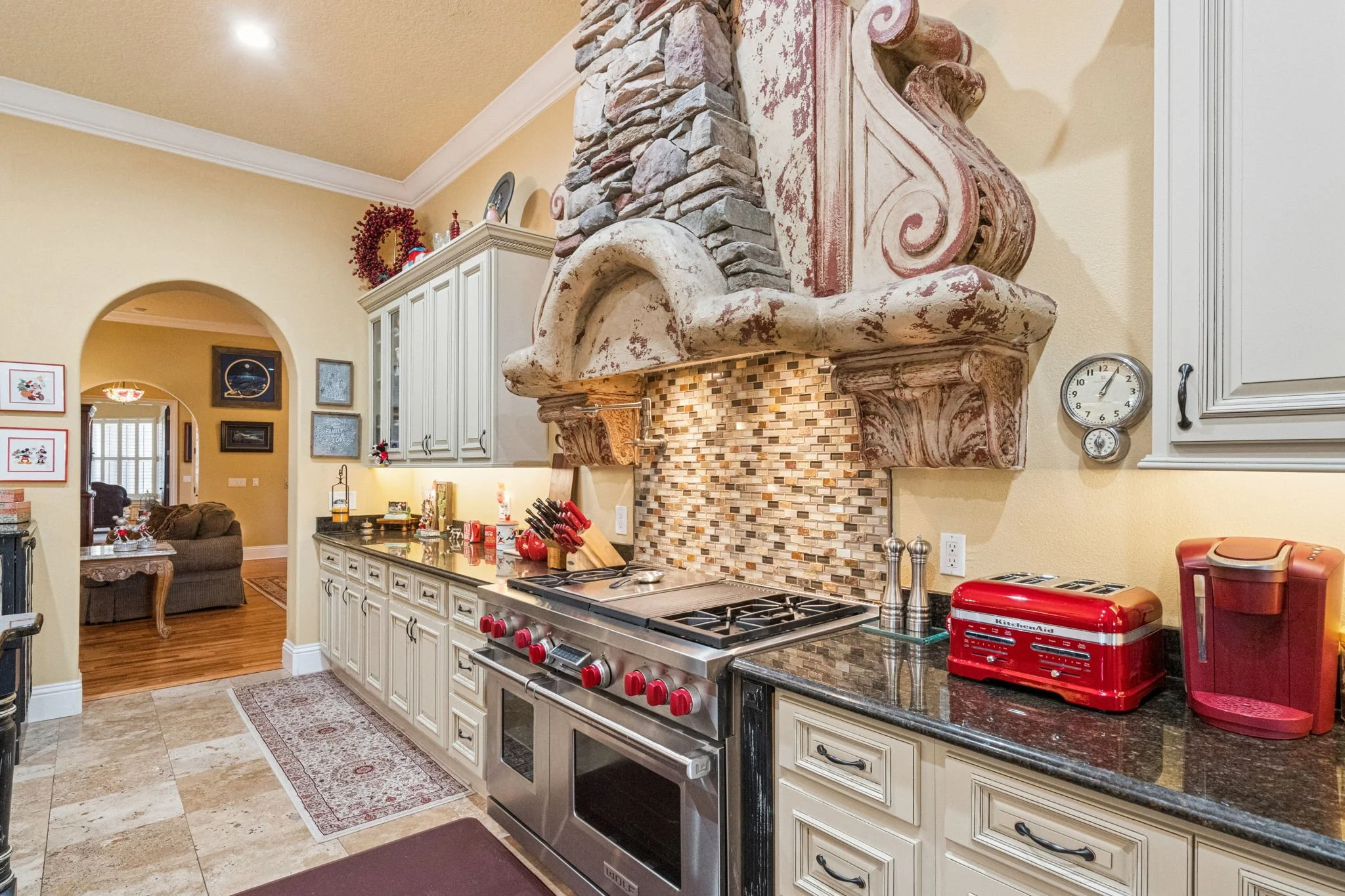 A kitchen with white cabinets, a decorative stone and wood range hood over a stainless steel stove, and various appliances on the countertop. A living room with a brown sofa and framed artwork is visible through an arched doorway.