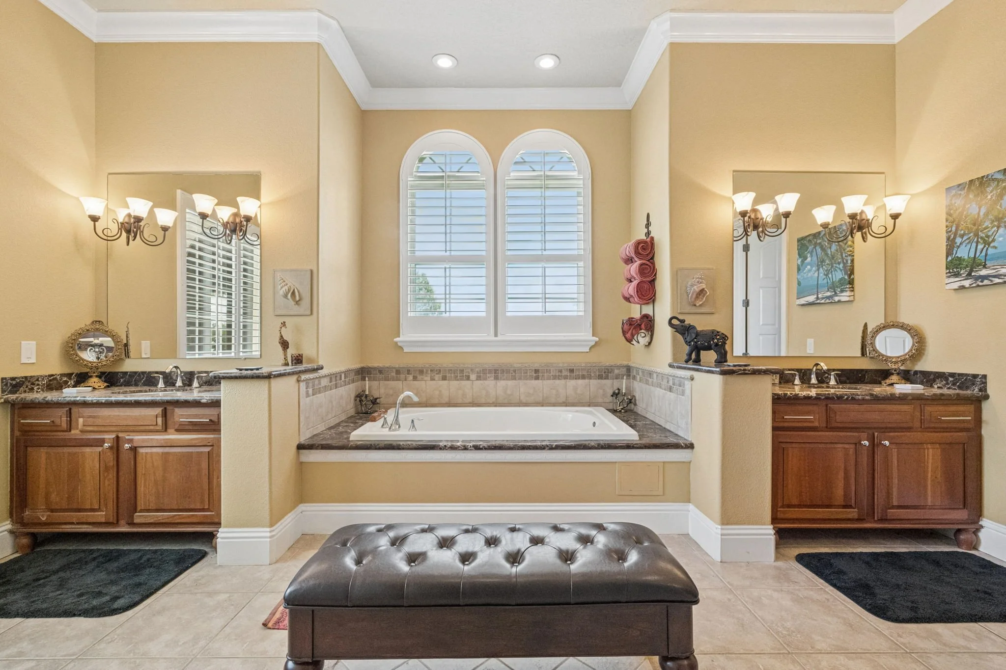 Bathroom with double sinks, large mirror, a bathtub between two windows, wooden cabinetry, wall sconces, and decorative accents.