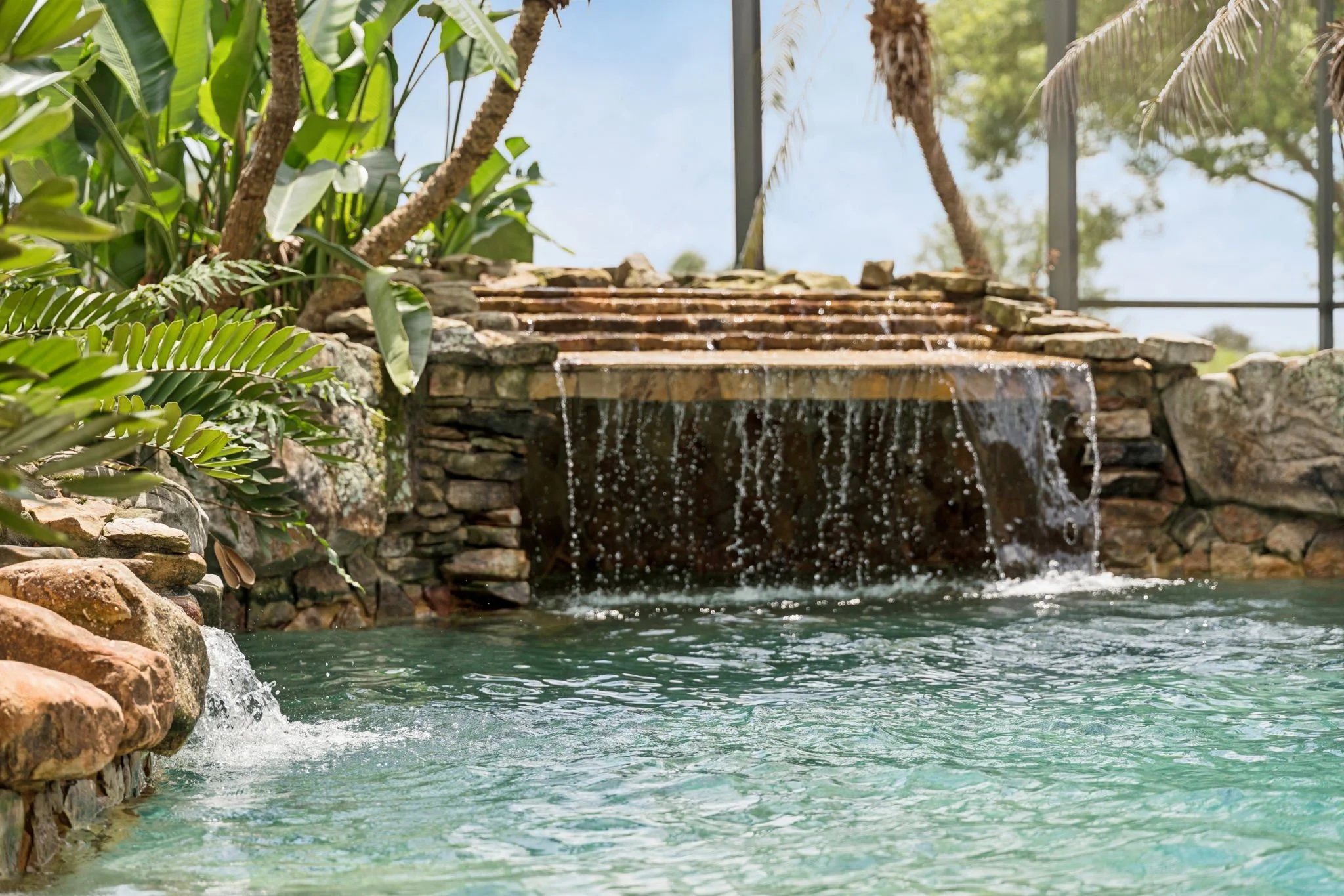 A small waterfall flows into a clear pool surrounded by rocks and tropical plants, with a screened enclosure and trees in the background.