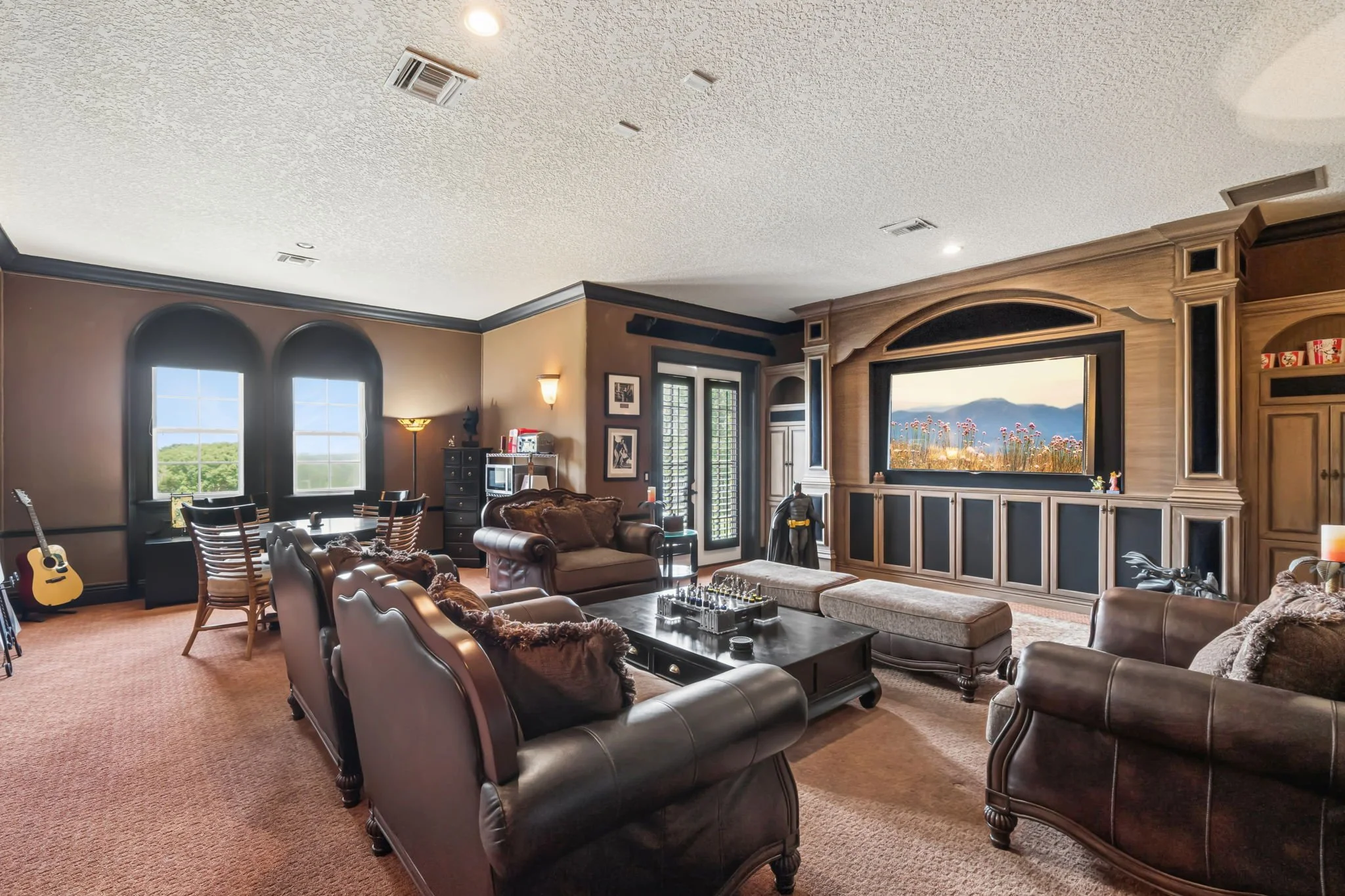 Living room with leather sofas, a coffee table, a music stand, a guitar near the window, artwork on the walls, a television showing a mountain landscape, and large windows letting in natural light.