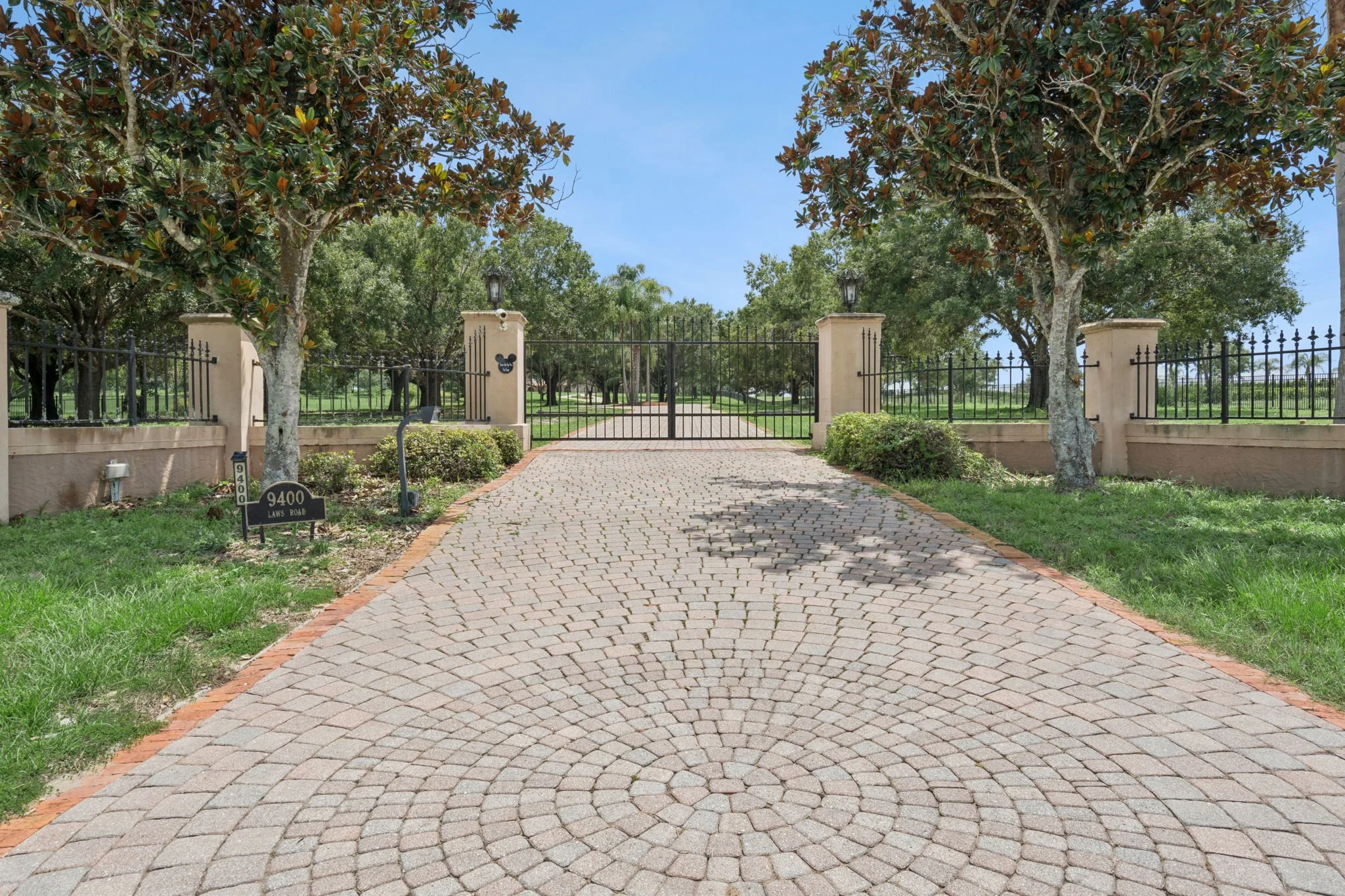 View of a gated entrance with cobblestone driveway, surrounded by trees and greenery, under a blue sky.