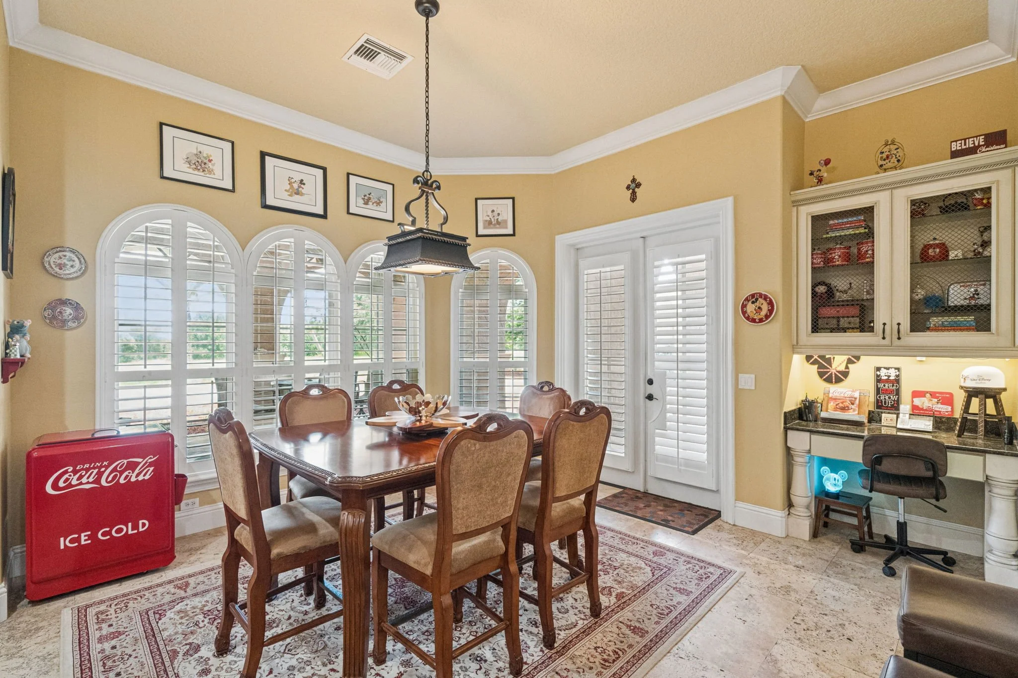 A dining room with a wooden table and chairs, large windows with shutters, wall decoration, a chandelier, and a corner with a kitchen countertop, cabinets, and a red Coca-Cola ice cooler.