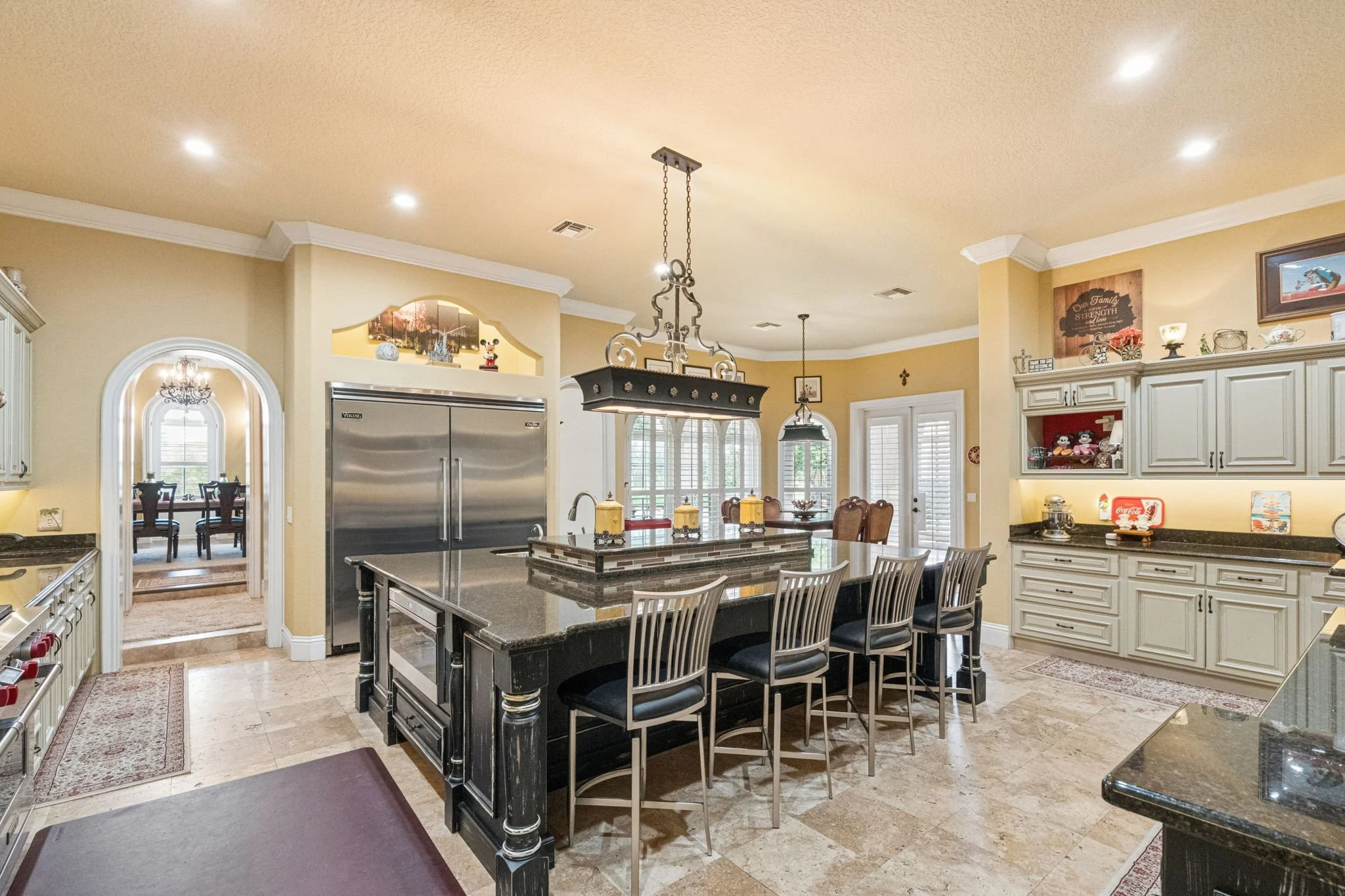A spacious kitchen with a central black island, white cabinetry, a stainless steel refrigerator, and decorative items on top of the cabinets. There is a dining area with chairs and a view of an adjoining room through an arched doorway. Recessed light