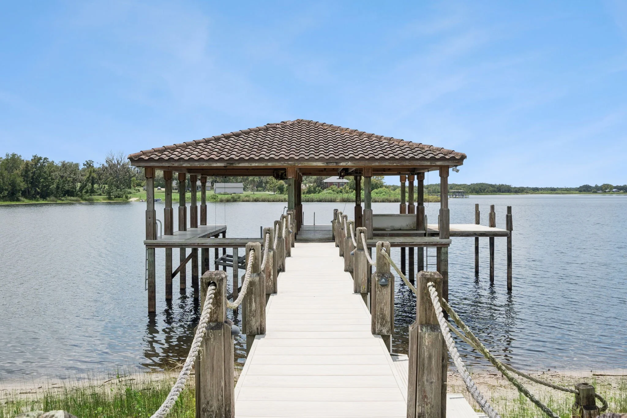 Wooden dock extending into a lake with a covered seating area at the end, surrounded by water and lush greenery, under a clear blue sky.