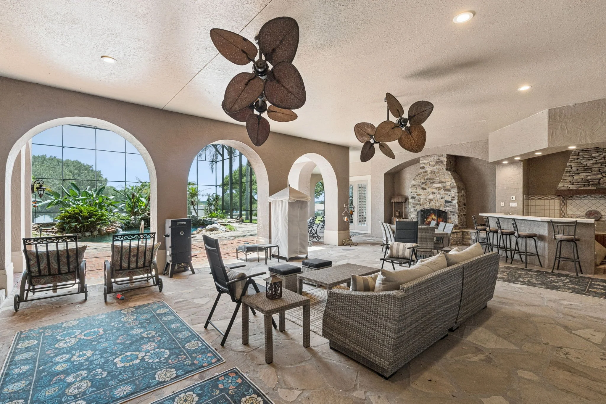 Indoor living area with large arched windows, ceiling fans with leaf-shaped blades, stone fireplace, bar area with high chairs, and outdoor patio with seating and greenery visible through the windows.