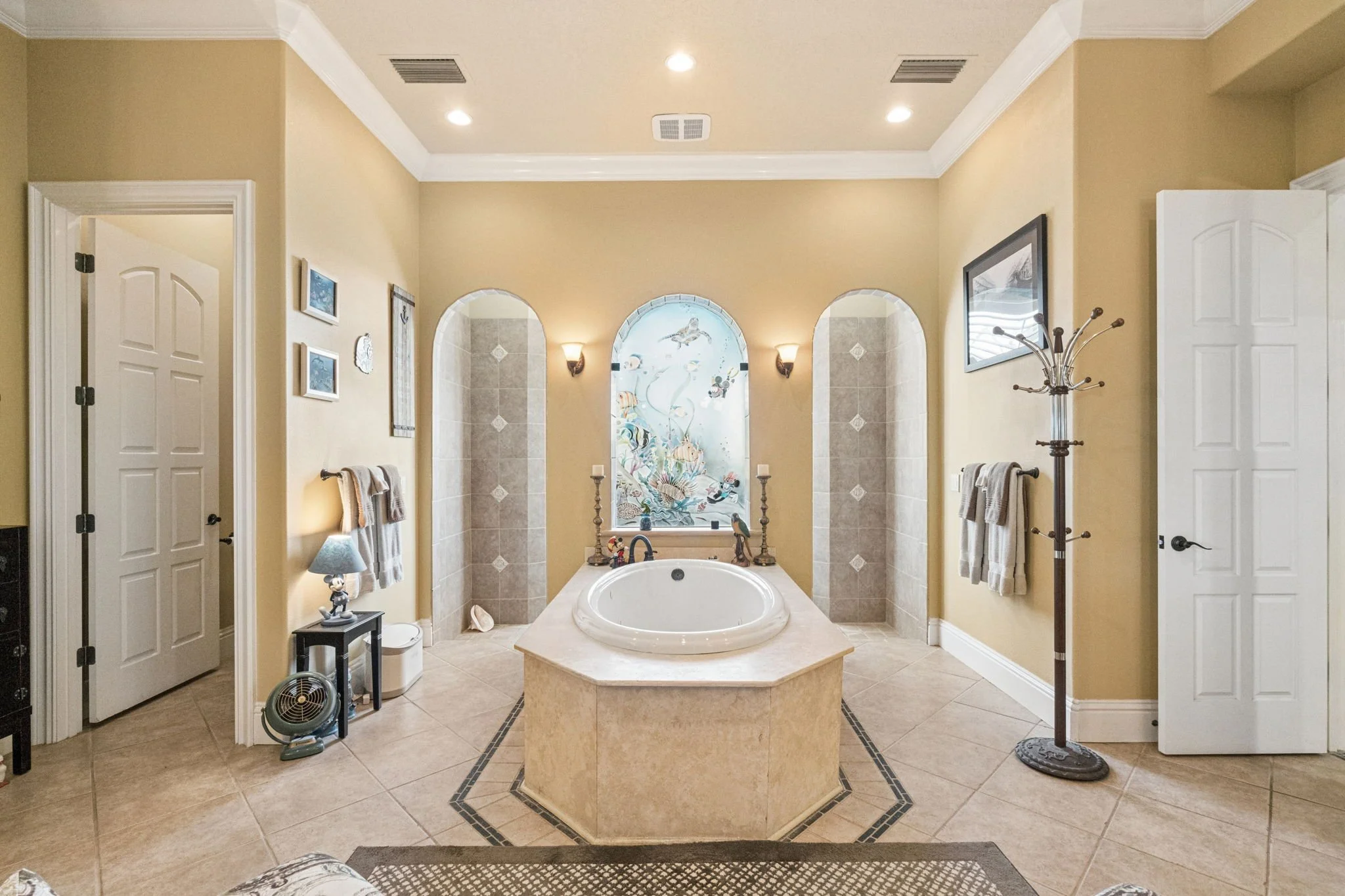 A luxurious bathroom with a large oval bathtub set in a tiled surround, a decorative stained glass window with fish art, surrounded by beige walls, white crown molding, and towels hanging on towel bars, with additional decorative items and lighting.