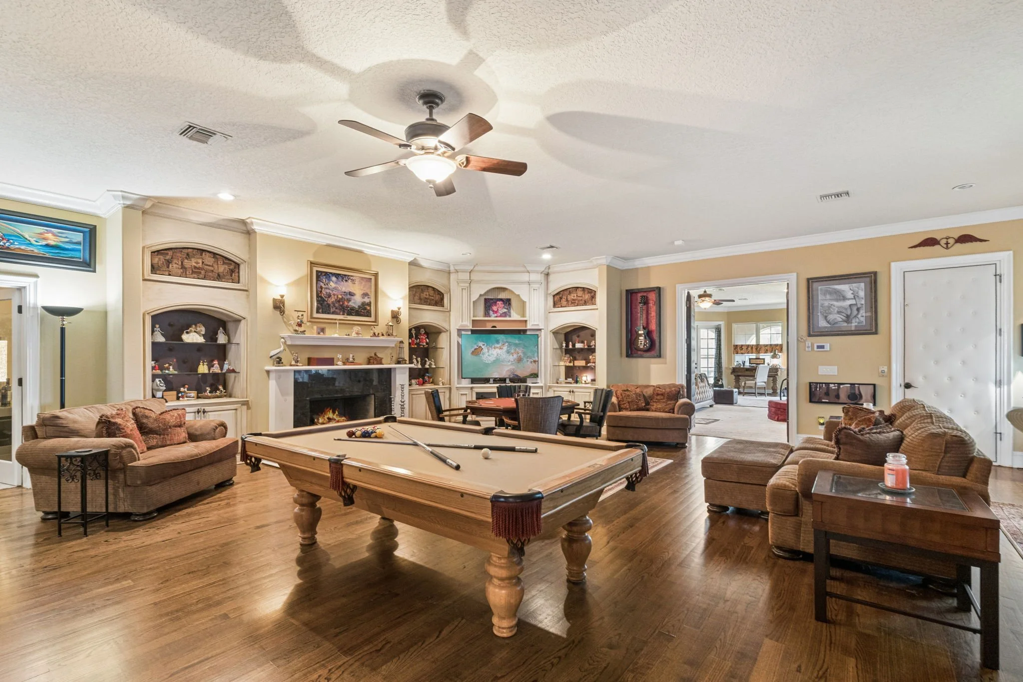 A spacious living room with a pool table, several sofas, a fireplace, wall decorations, a television, wooden flooring, and a ceiling fan.