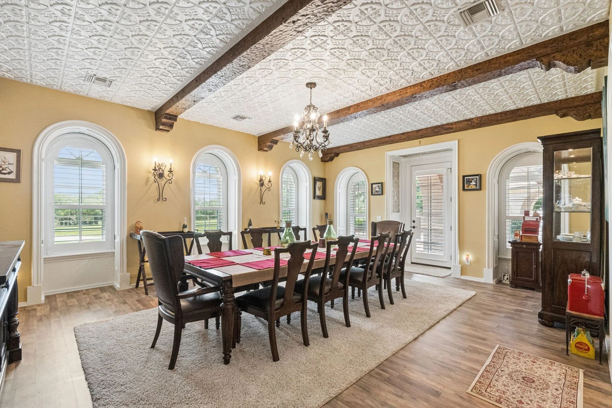 A dining room with a long wooden table covered with a red tablecloth and surrounded by dark wooden chairs. The room has yellow walls with decorative wall sconces, arched windows with shutters, and a ceiling with intricate white tin tiles and dark woo