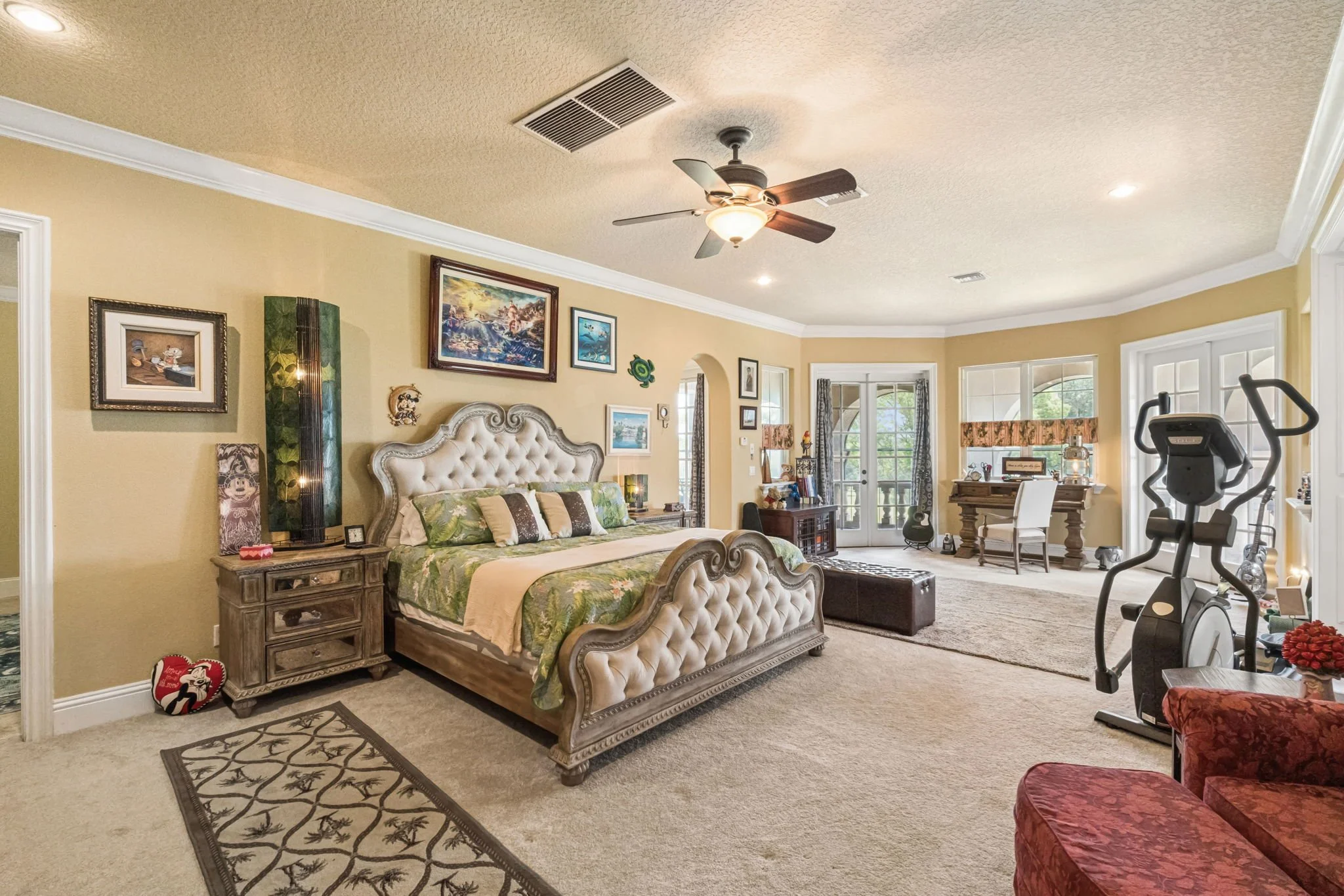 A spacious, sunlit living room with a large bed featuring an ornate wooden frame, a collection of framed pictures and artworks on the beige walls, an exercise machine in the corner, a dining table with chairs near large windows, and a ceiling fan wit