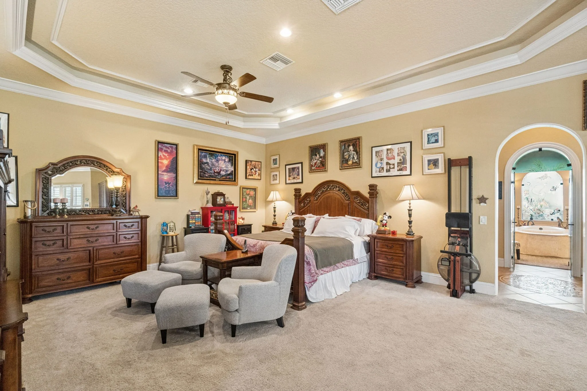 A luxurious bedroom with beige walls and elaborate white crown molding. The room has a wooden bed with a carved headboard, covered in white bedding with pink accents. There are two desks, one with a large mirror, a small red bookshelf, and a beige ar