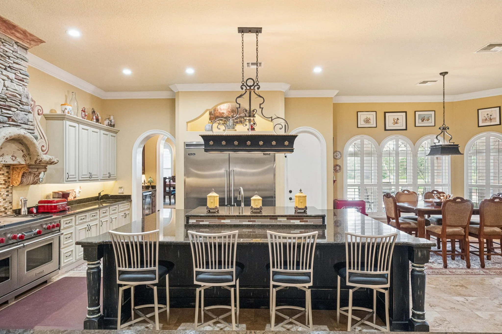 A spacious kitchen with a central island, white cabinets, stainless steel appliances, and a dining area with upholstered chairs and large arched windows.
