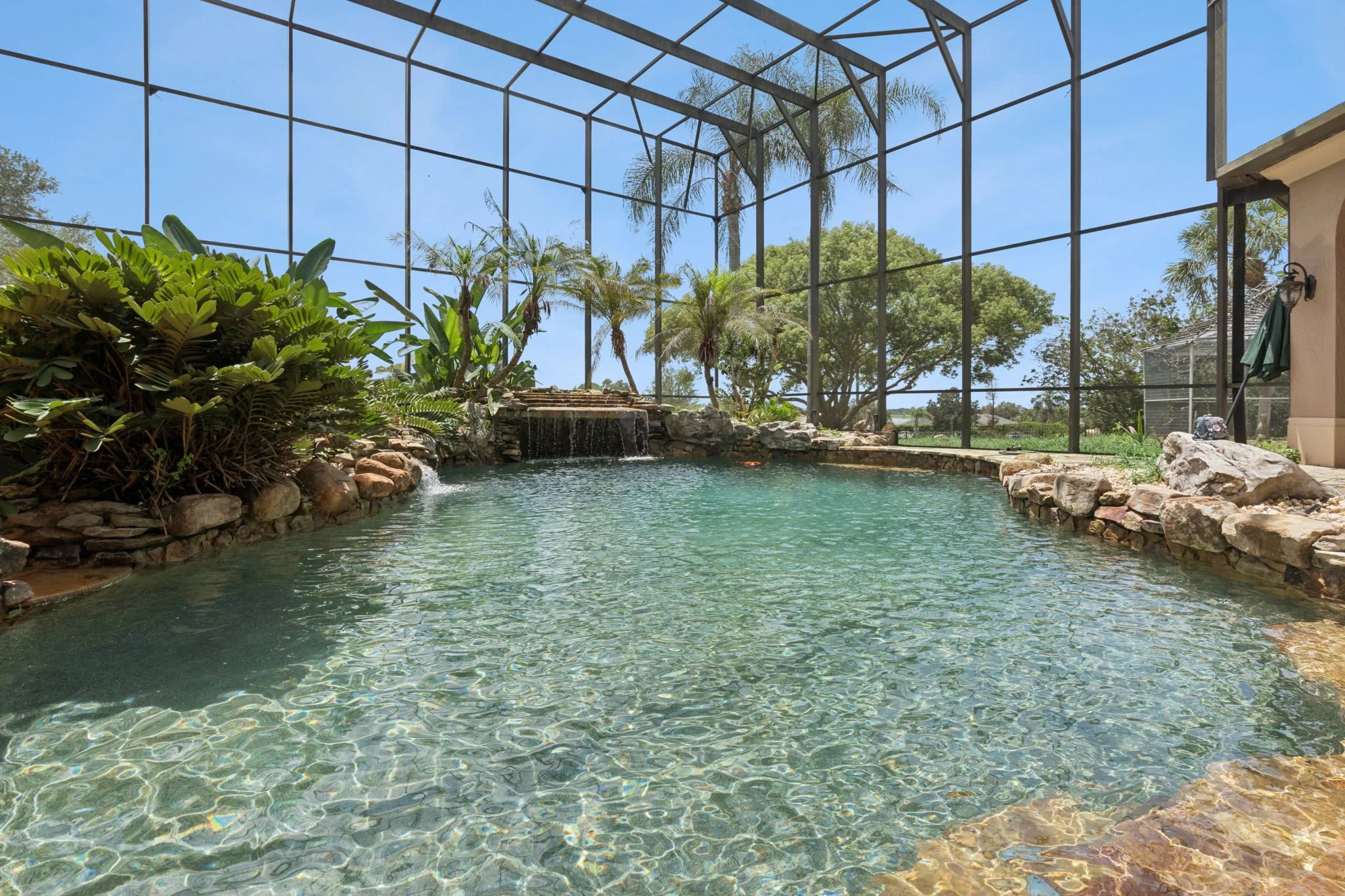 Indoor swimming pool with a waterfall feature, surrounded by rocks, with tropical plants and trees visible through a screened enclosure and a clear blue sky outside.