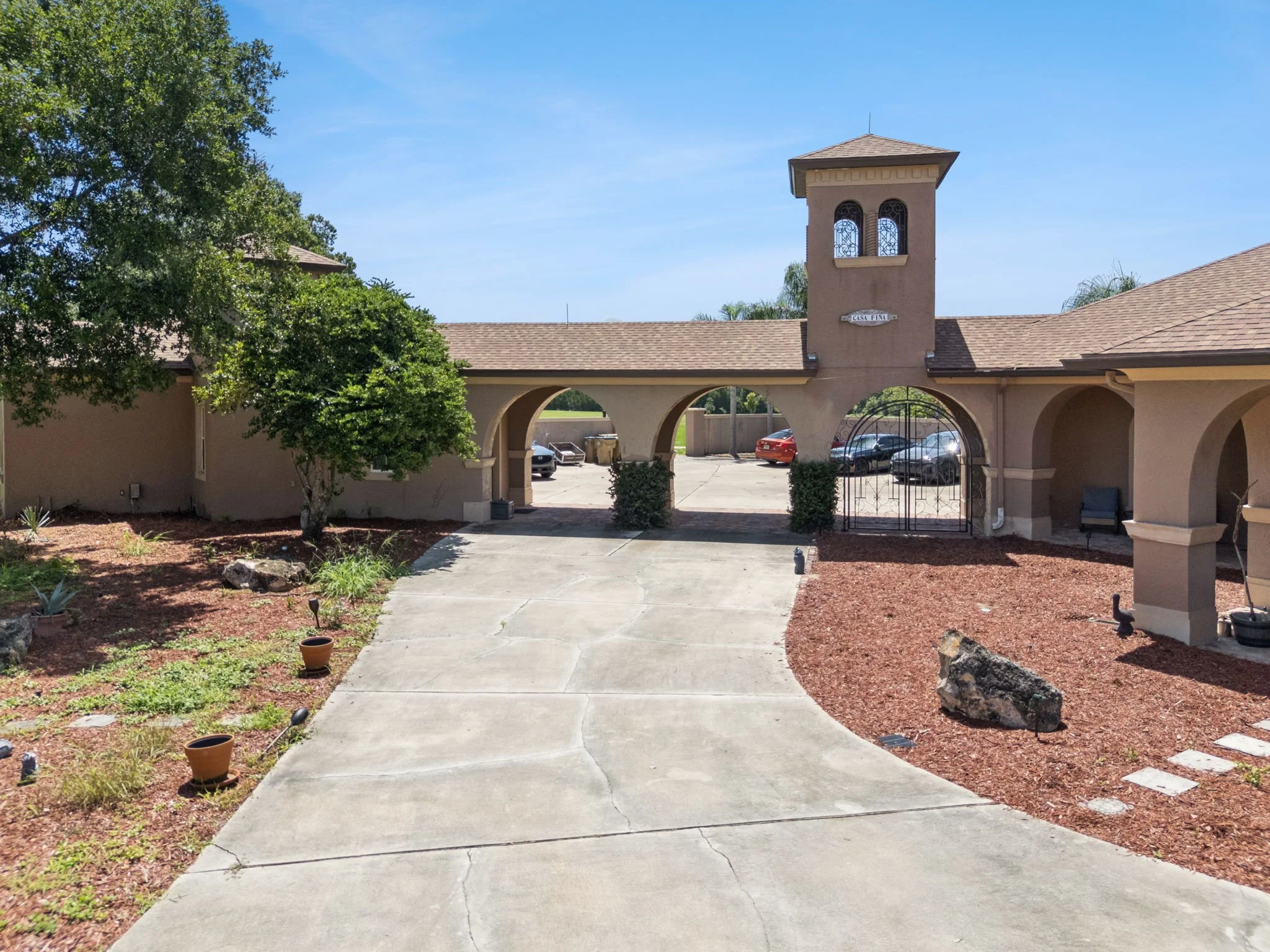 View of a building with a tower and an arched entrance, surrounded by trees and a parking lot with cars, under a clear blue sky.