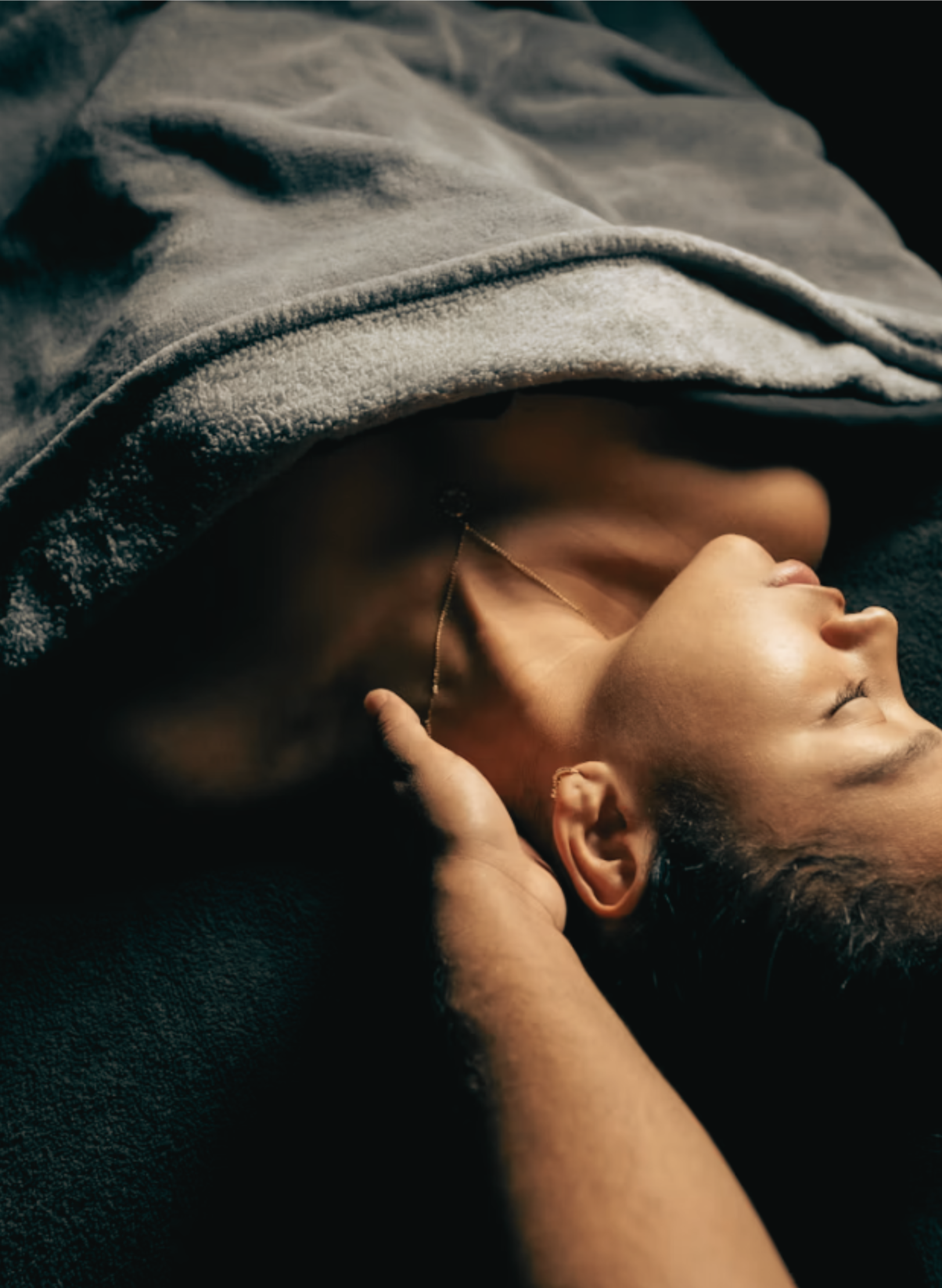 A person lying down with a hoodie over their face, wearing a necklace, with hands gently touching their head and face, on a dark surface.