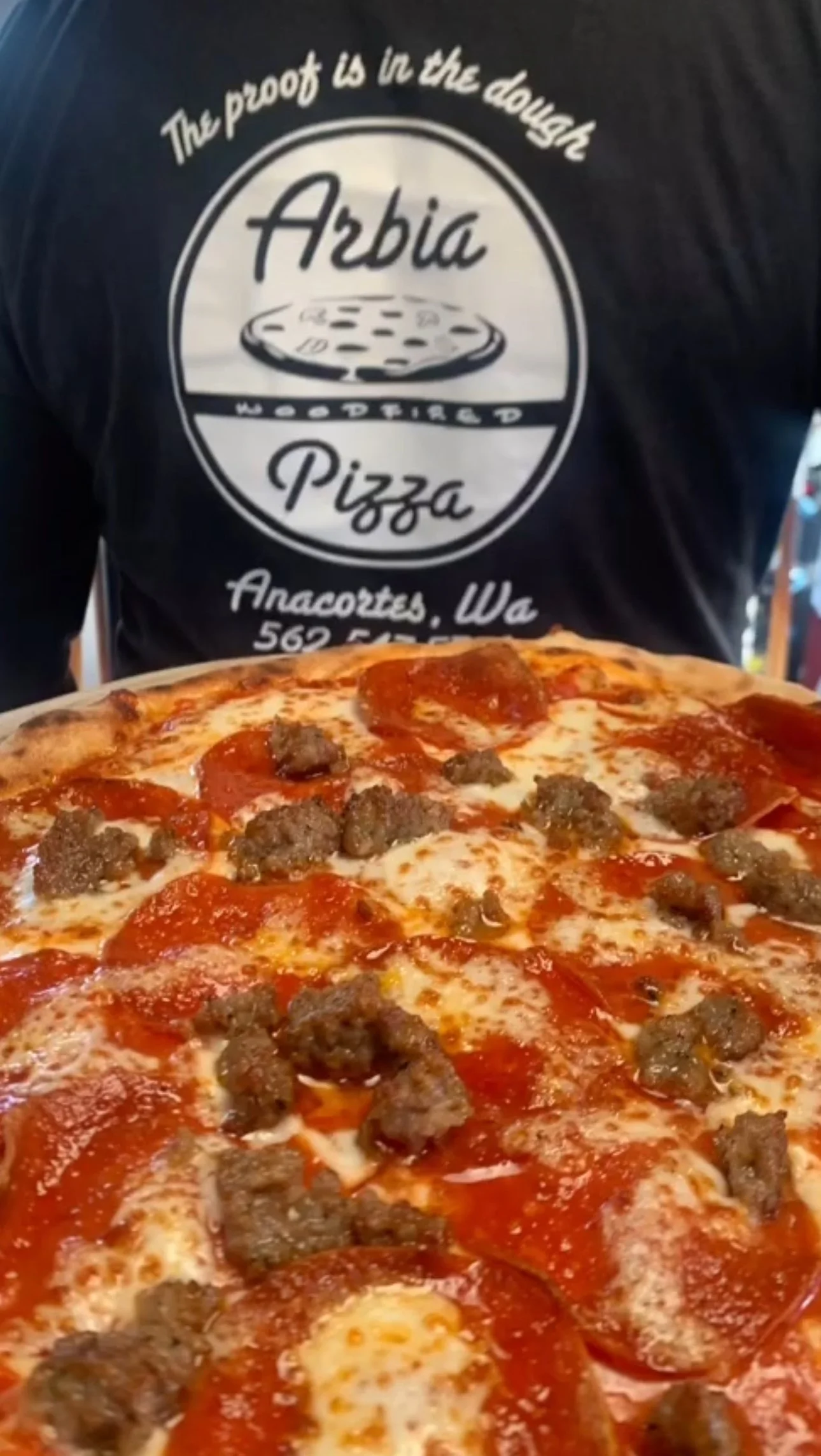 Close-up of a pepperoni and sausage pizza in front of a person wearing a black shirt with a logo for Arbia, a mobile pizza business in Anacortes, Washington.