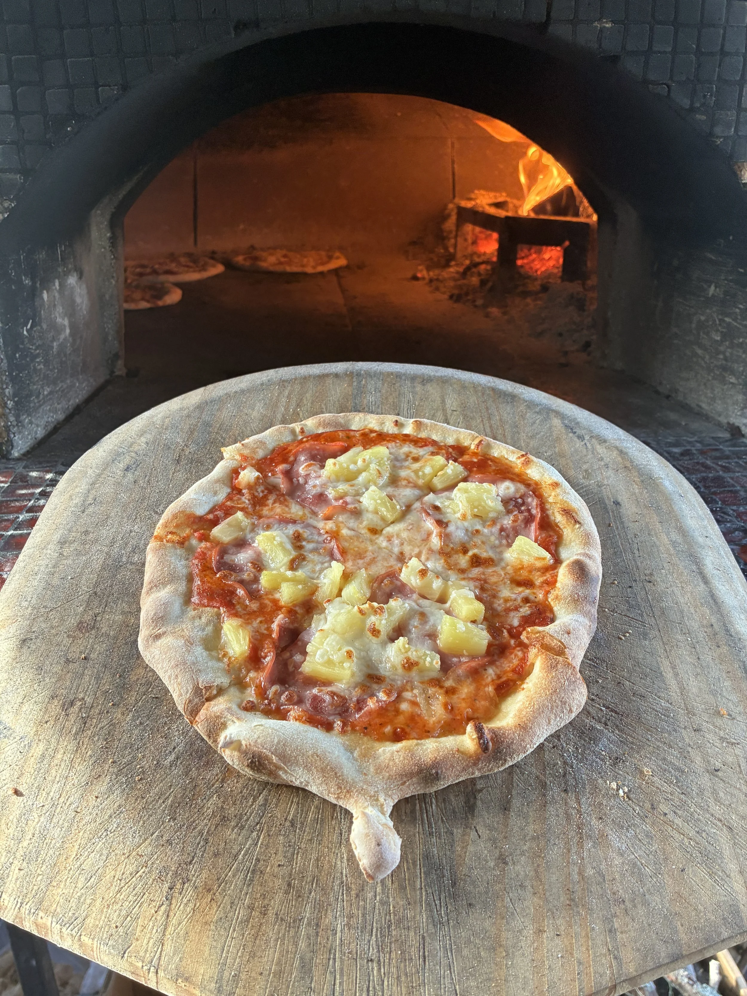 Pizza in a wood-fired oven with a crust, cheese, ham, and pineapple topping on a wooden pizza peel.