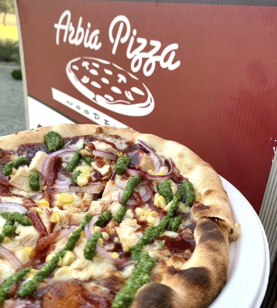 A pizza topped with corn, onions, chicken, and green pesto sauce.