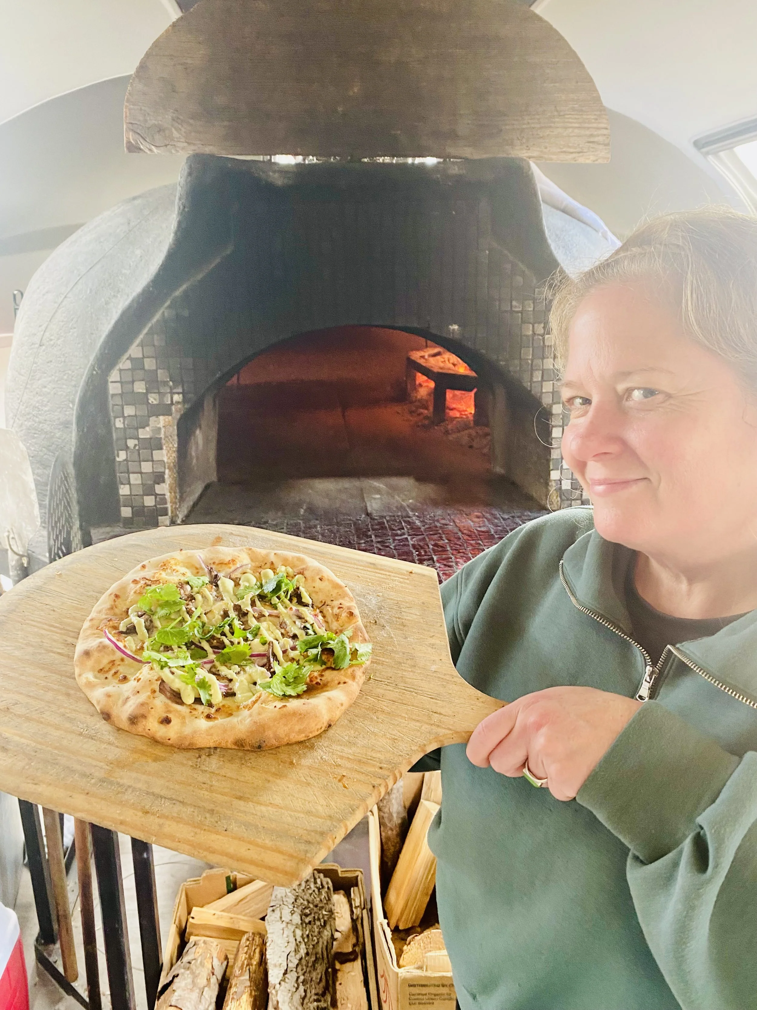 A woman holding a pizza on a wooden pizza board in front of a wood-fired oven with logs stacked nearby.