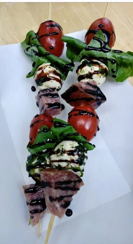 Skewers with cherry tomatoes, mozzarella balls, basil leaves, and grilled meat, topped with balsamic glaze and chocolate drizzle.