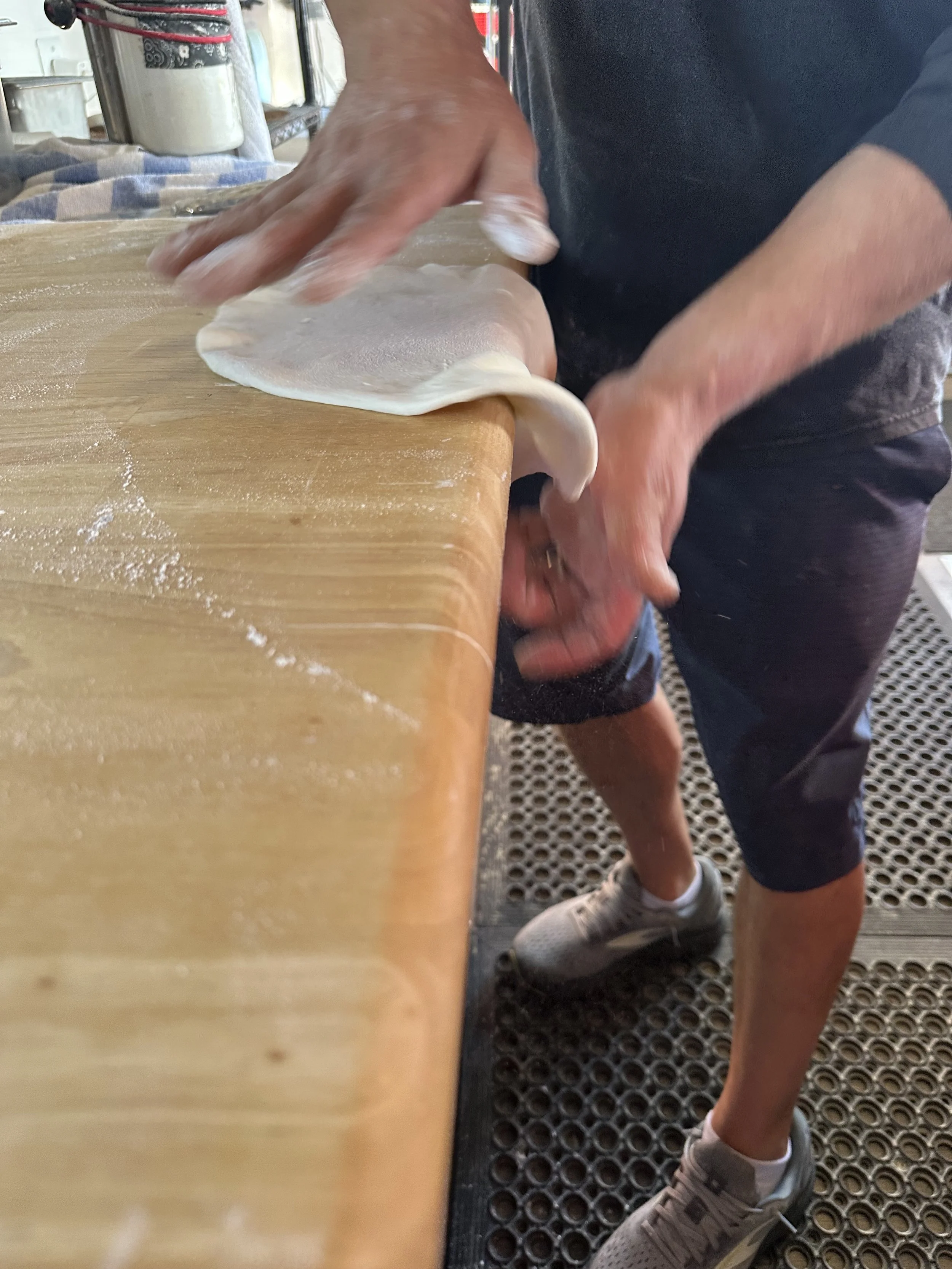 Person working with dough on a wooden surface, wearing sneakers and shorts, in a bakery or kitchen setting.
